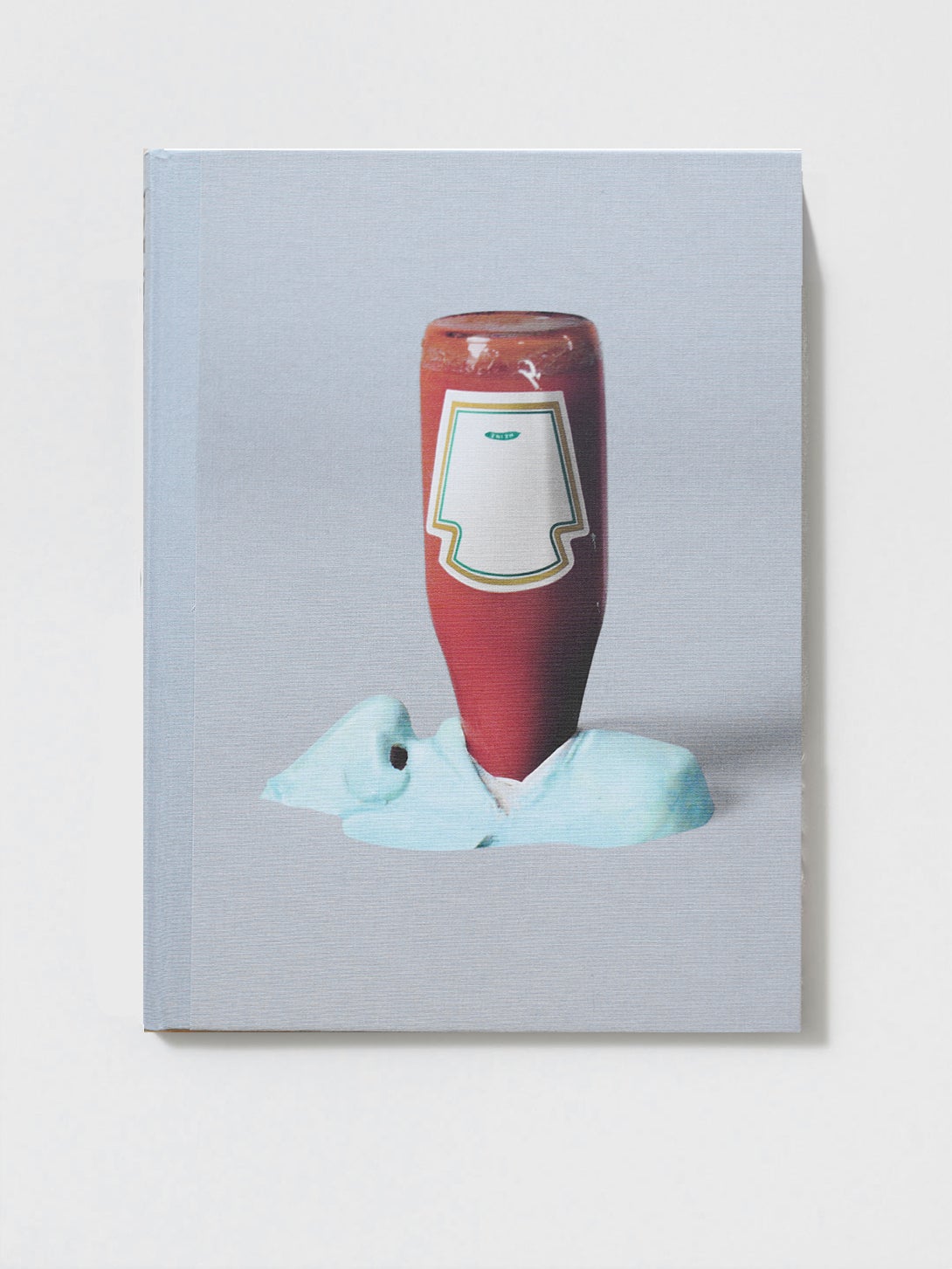 A book sits on a white surface, the cover has a blue mouth with a bottle of ketchup in it.