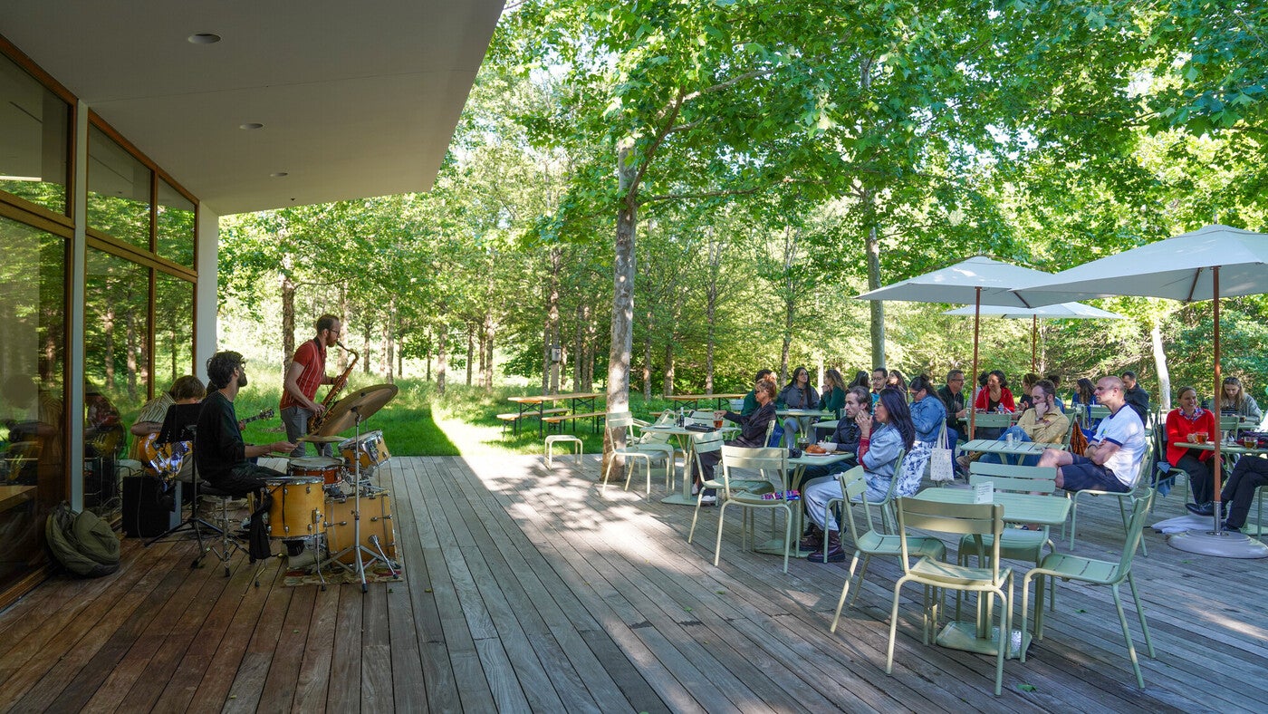 People at outdoor tables amidst green scenery listening to a small band near a building