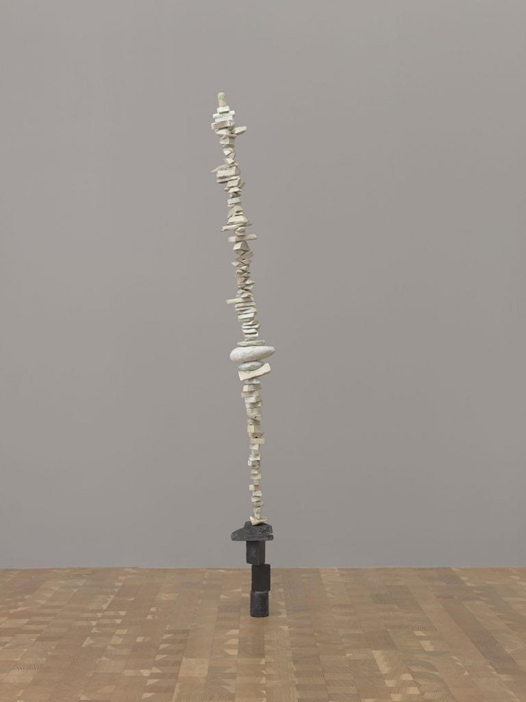 Una delgada escultura compuesta por bloques de madera apilados pintados de blanco, con una base de madera pintada de negro, se inclina ligeramente hacia la izquierda en la galería.