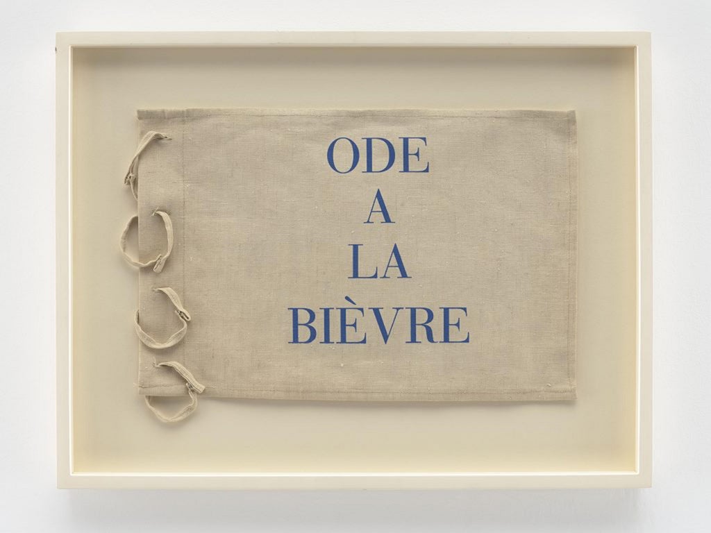 A beige cotton-looking rectangle with the words Ode de La Bievre printed in blue capital letters in the center vertically. On the left side of the rectangle are four attached vertical cotton loops.