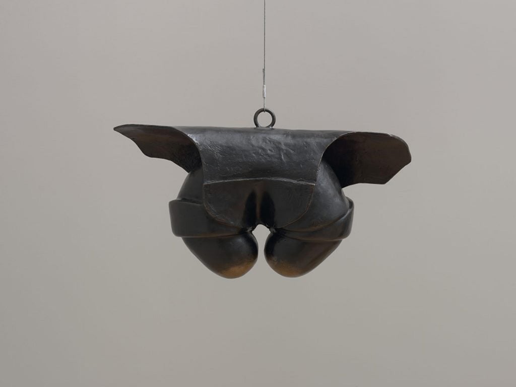 Un cable vertical cuelga de la parte superior central, conectado a un pequeño círculo del que cuelgan formas horizontales en forma de alas Debajo de estos hay tocones cortos y redondeados que miran hacia adentro.