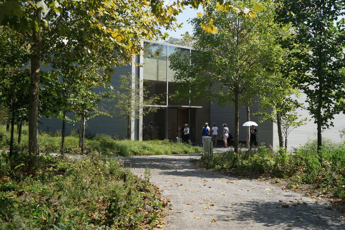 A pathway in the foreground leads to a building (the Arrival Hall) with large glass windows and doors. A group of people approach the building.