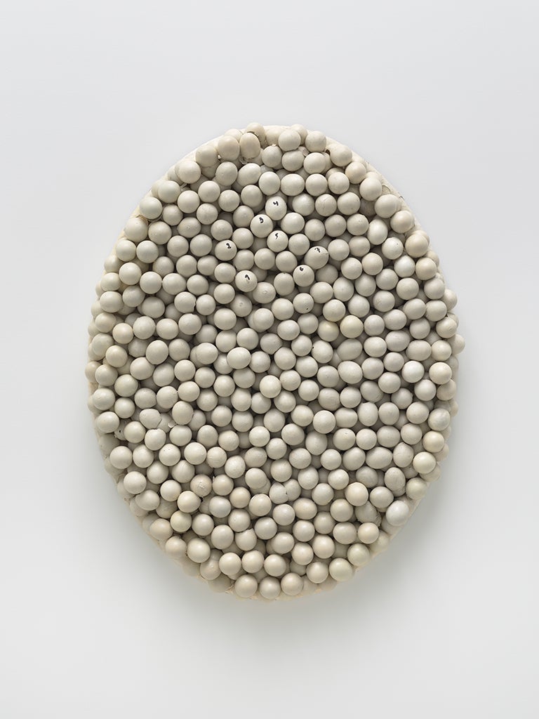 An oval sculpture covered by unbroken eggshells sits on a wall. Seven eggs are labeled one through seven in black marker.