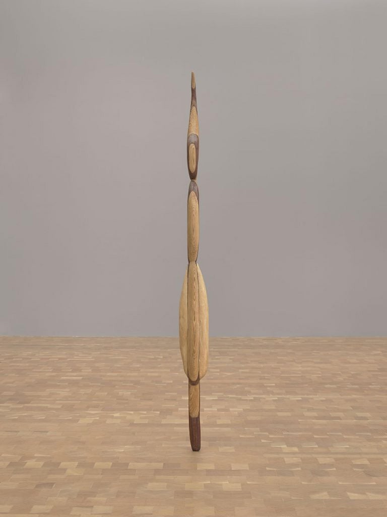 Una escultura de madera segmentada alta, delgada, de color beige y marrón rojizo, está adherida al piso de patrón cuadrado frente a una pared pintada de gris.