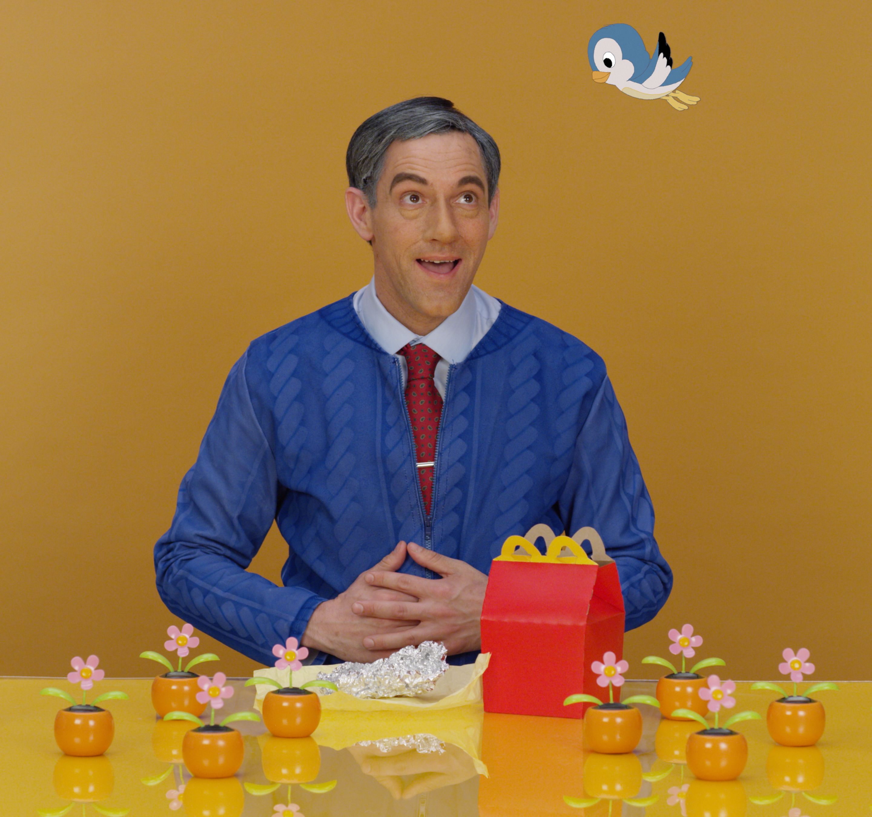 A man looks up at a cartoon bird andsits at a table littered in plastic toys and a McDonalds Happy Meal box