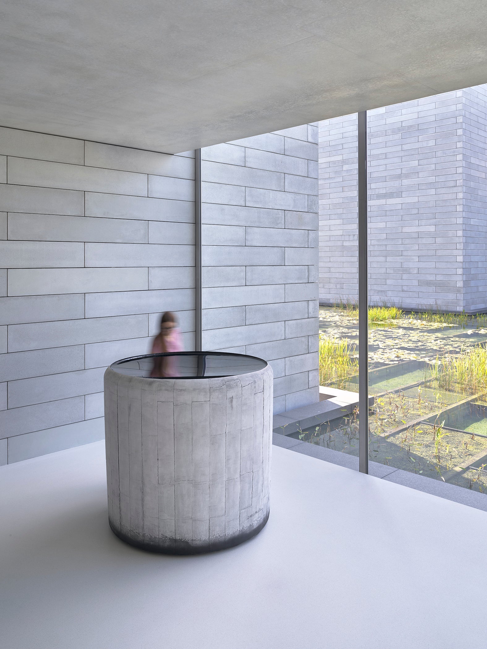 Blurry figure of a person stands next to glass cylinder and window overlooking a water court