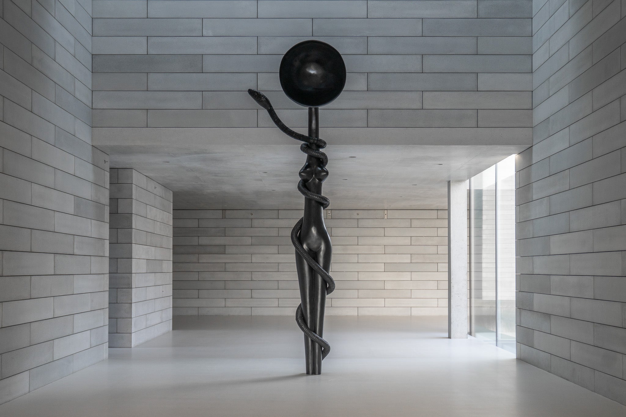 A large bronze female form wrapped in a snake stands in a concrete room with windows.