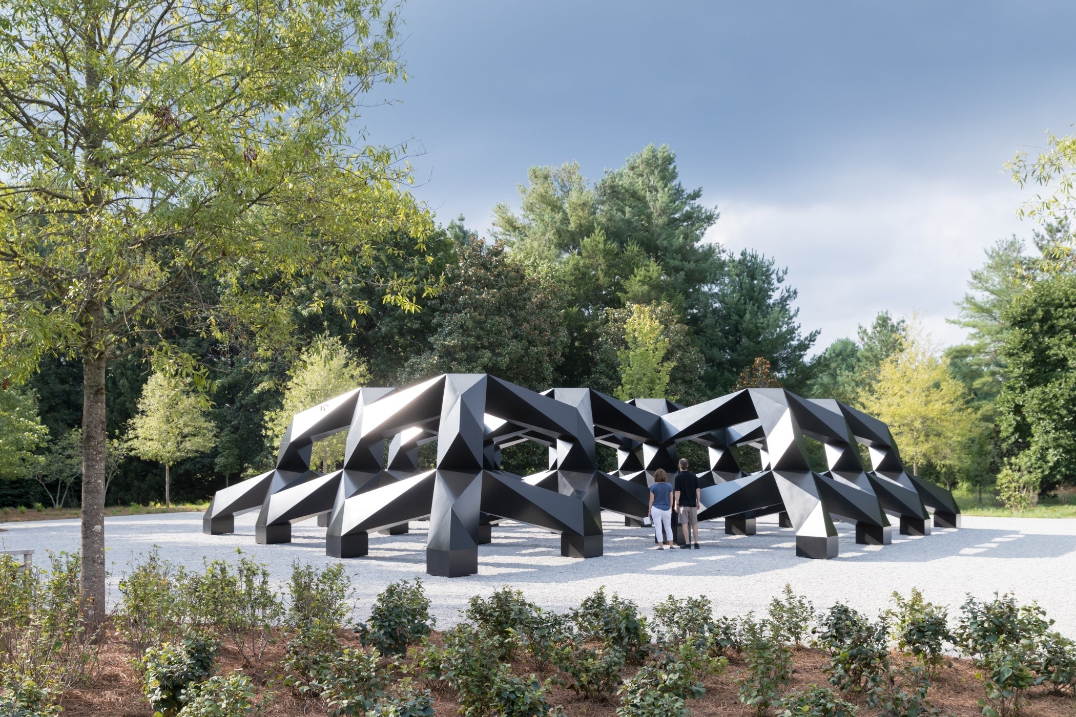 Large geometric sculpture sits amidst gravel and foliage