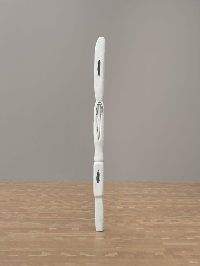 En la galería hay una escultura de madera blanca pintada, alta, delgada y de forma segmentada, como un tótem, con contornos negros.