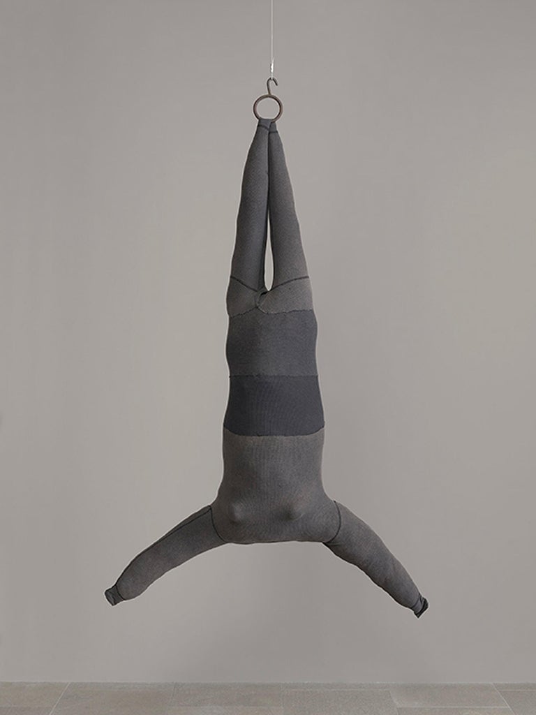 Una figura de tela extendida sin cabeza segmentada de tamaño completo cuelga "al revés", suspendida del techo con un cordón que se sujeta a un gancho y un anillo de soporte.