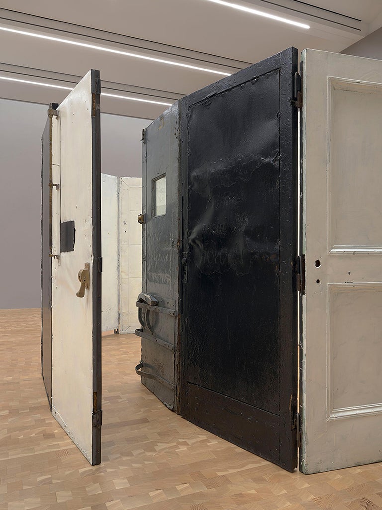 Una cerradura con puertas industriales batientes de madera pintada y acero. La habitación cuenta con una combinación de objetos surrealistas, ordinarios y extraordinarios de mármol, acero, vidrio y un espejo.