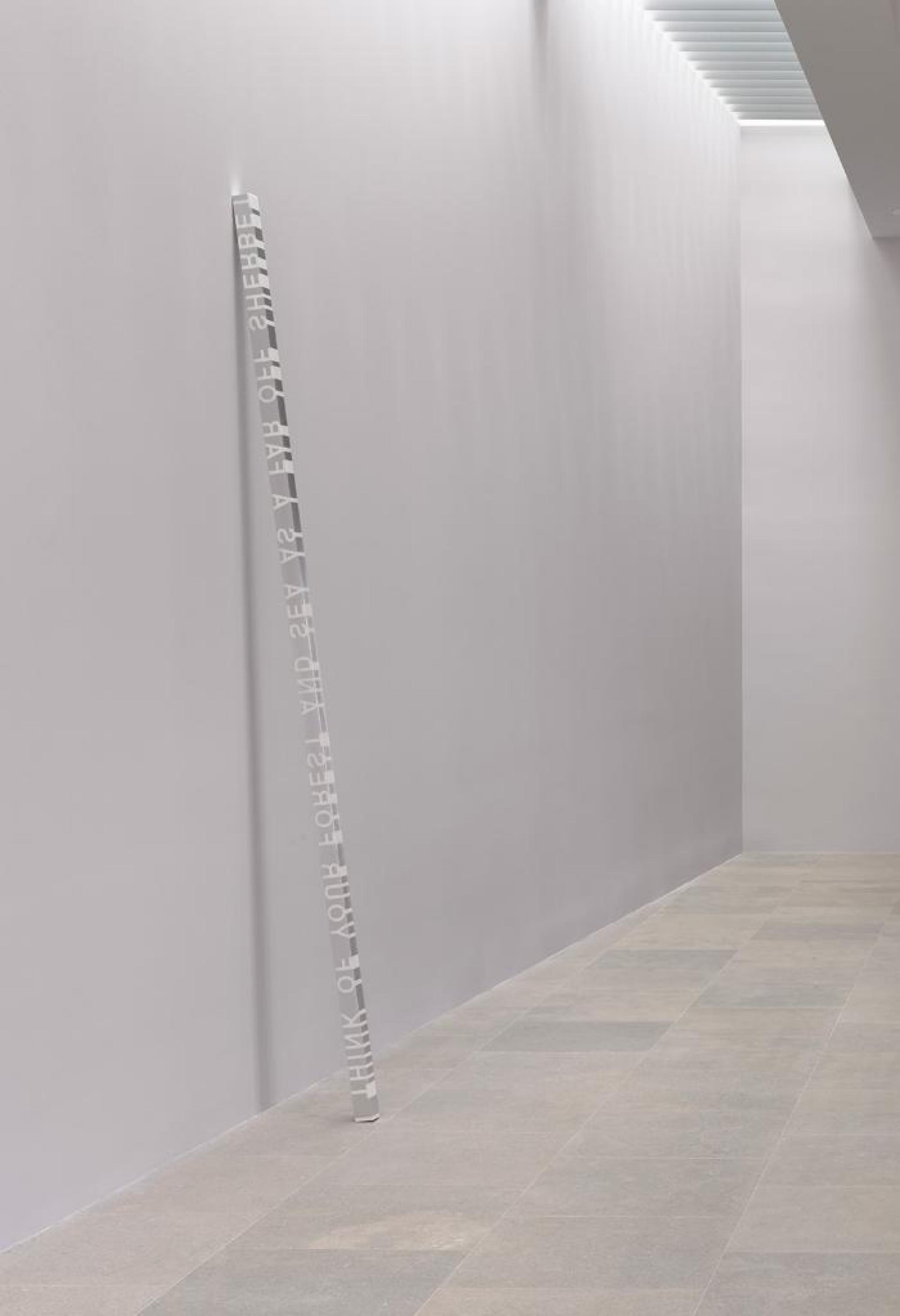 A clear column with I THINK OF YOUR FOREST AND SEA AS A FAR OFF SHERBET written inside leaning on a wall.