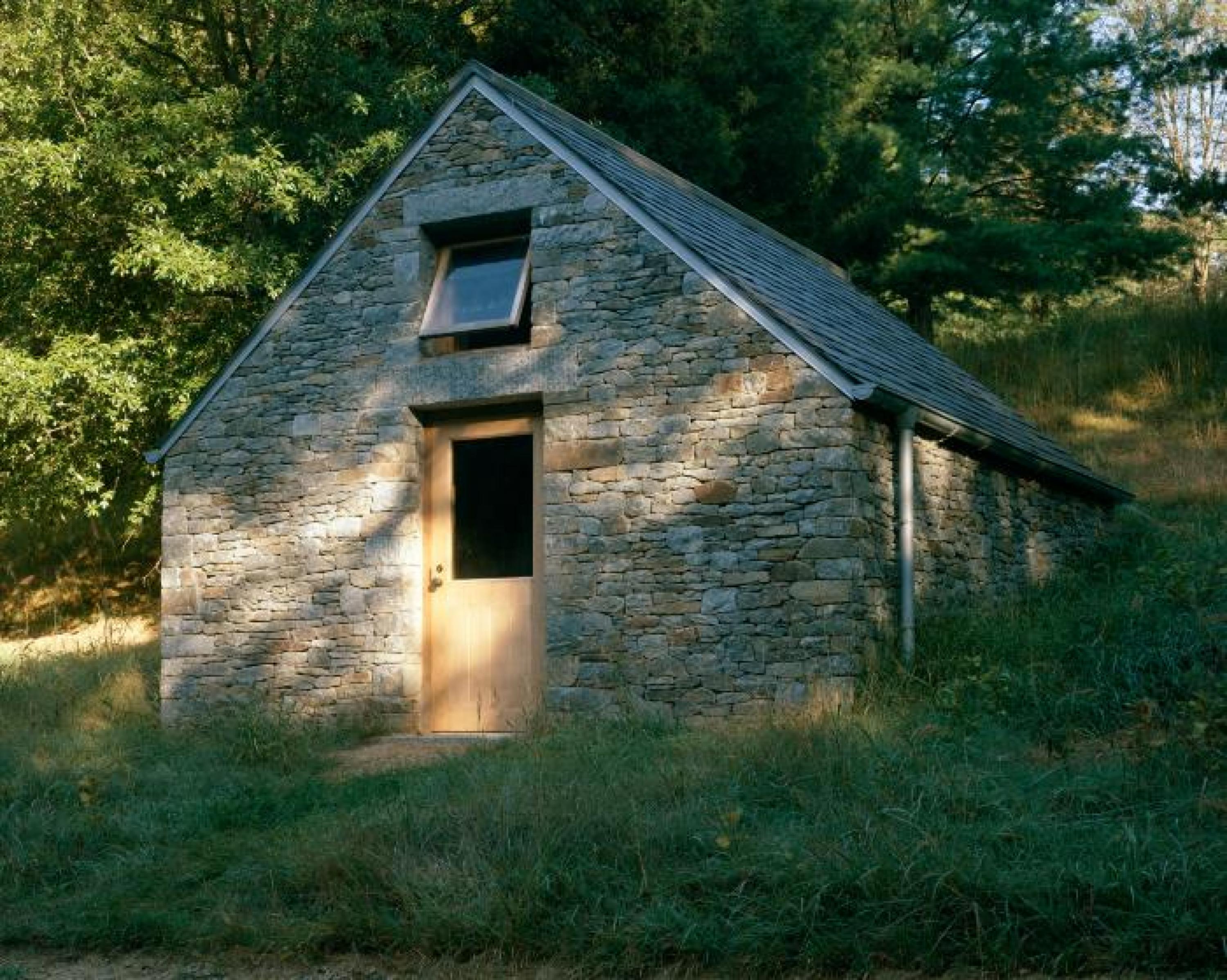A small stone house with a wooden door sits in a grassy hill. The window above the door is propped open.