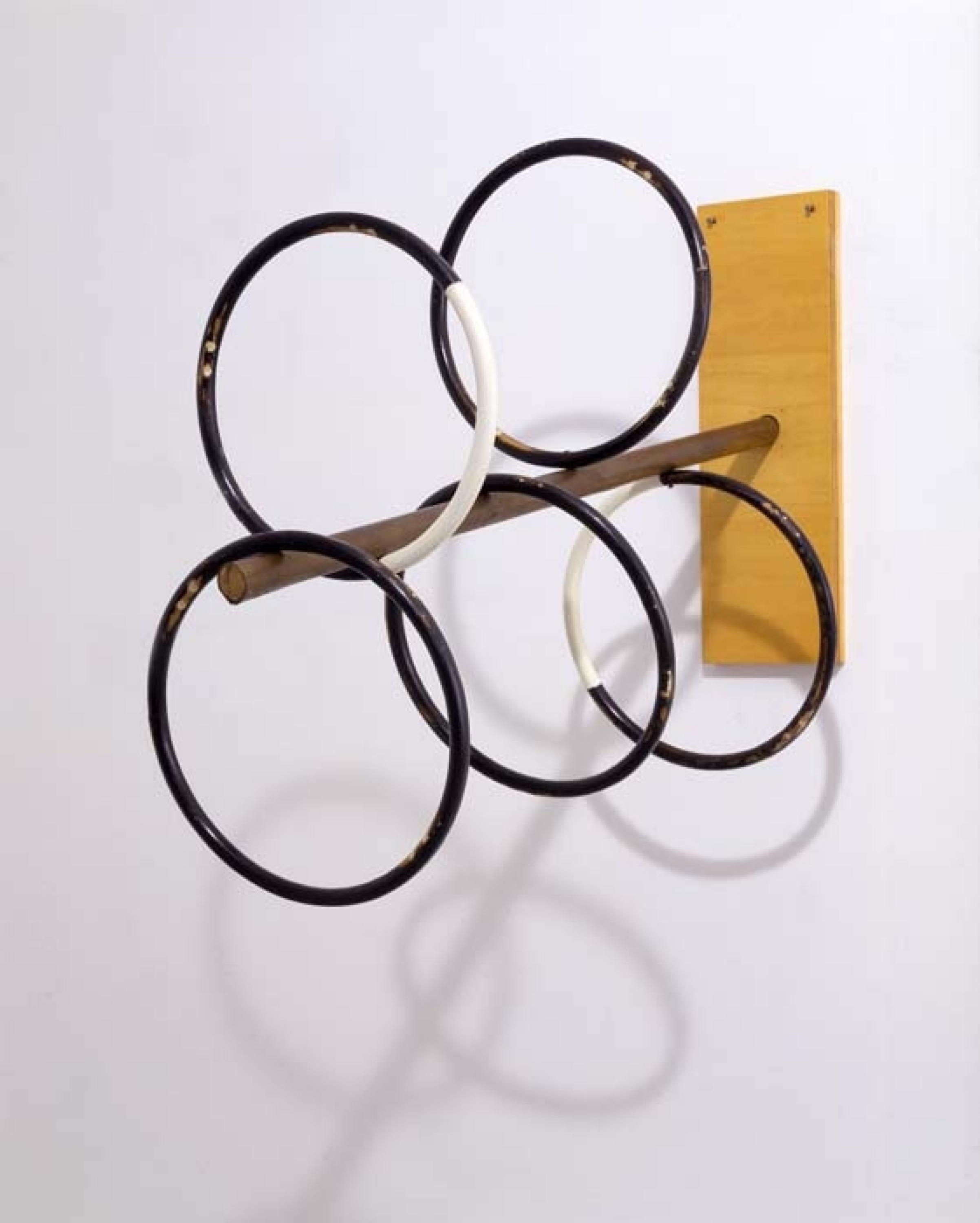 A scultpure juts out from a white wall. There is a rectangular base attached to the wall, a stick protrudes from the center of the base. There are five black and white circles attached to the stick.