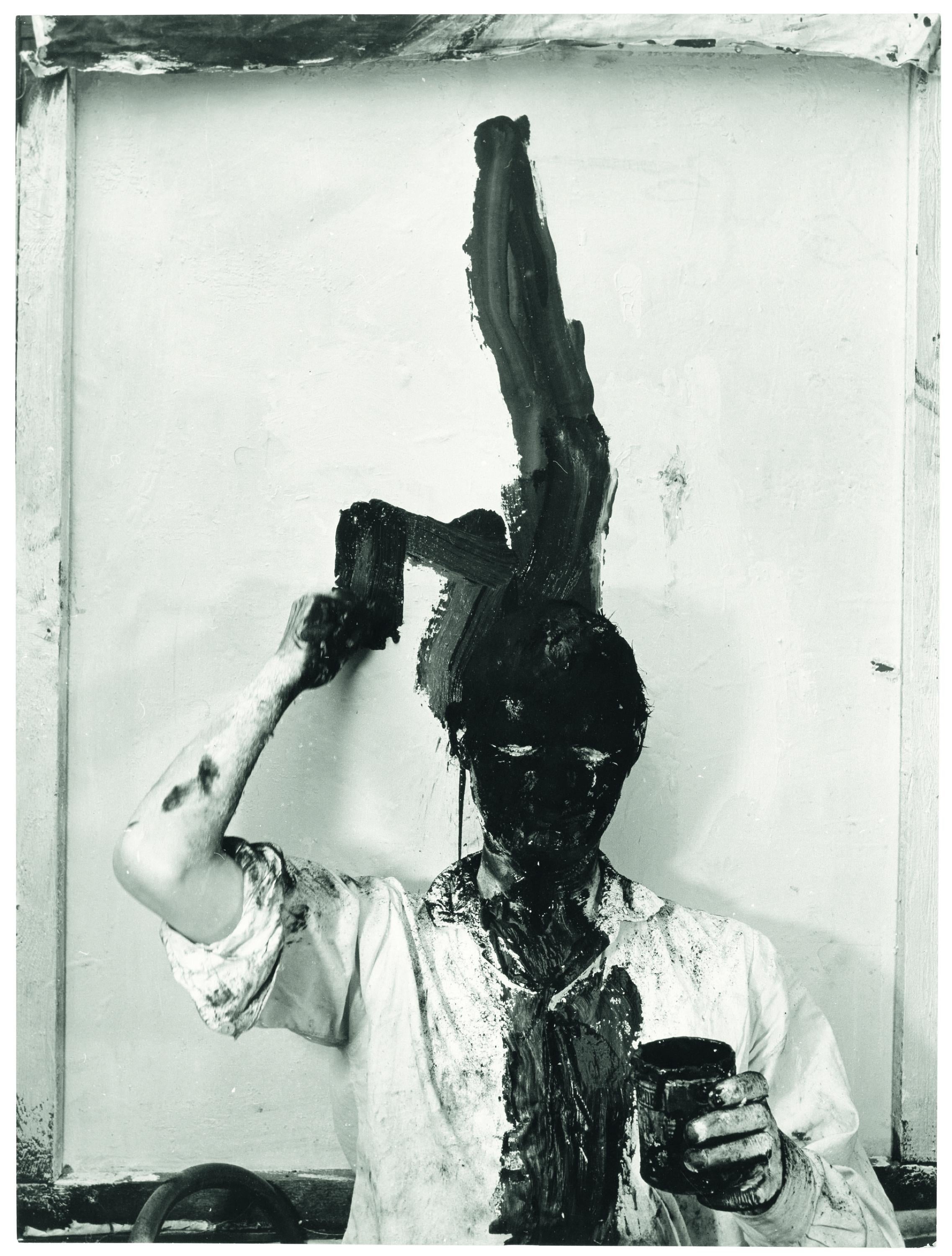 A black-and-white photograph of a man standing in front of a white wall, painting a thick black streak from above his head down across the center of his white shirt. His face is painted entirely black.