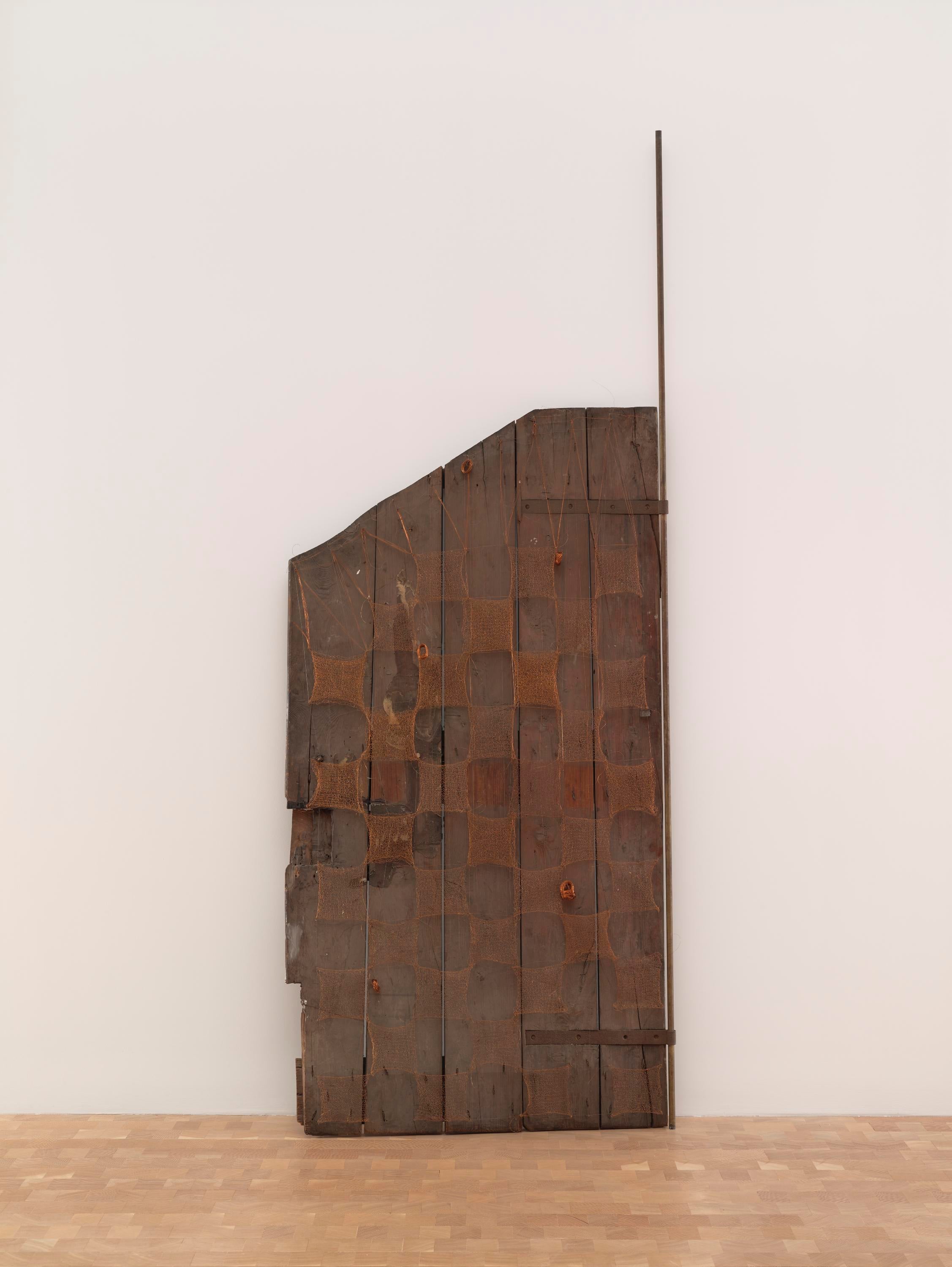 A wooden door leans against a wall. It is covered in squares of hand-woven copper wire and has a dusty patina.