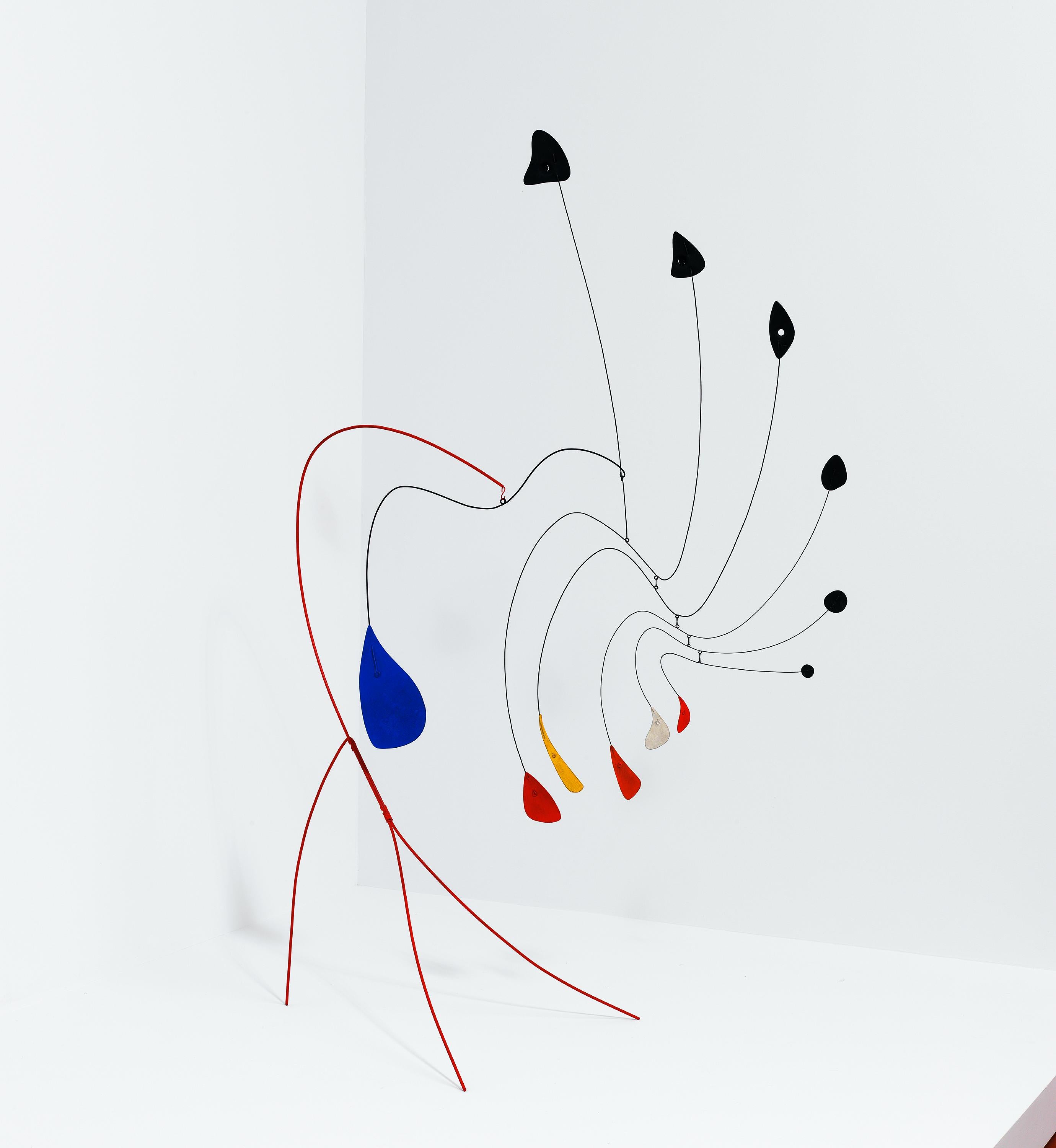 Hanging from a tripedal floor stand, a series of graduated abstract forms (painted black, blue, yellow, red, and white) delicately balance on thin wires.