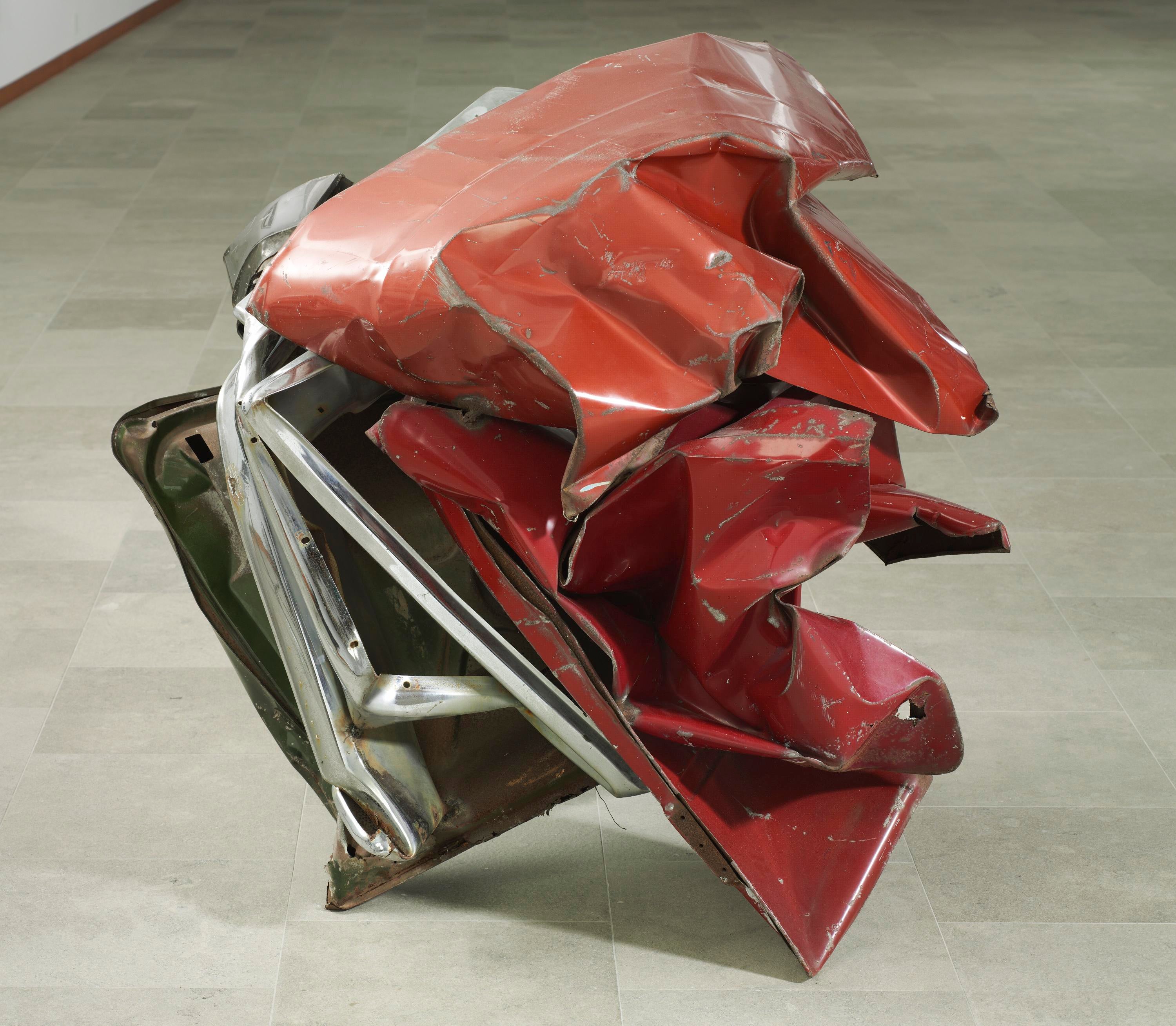 Crumbled like a piece of paper, car parts painted red balance on a slate floor.