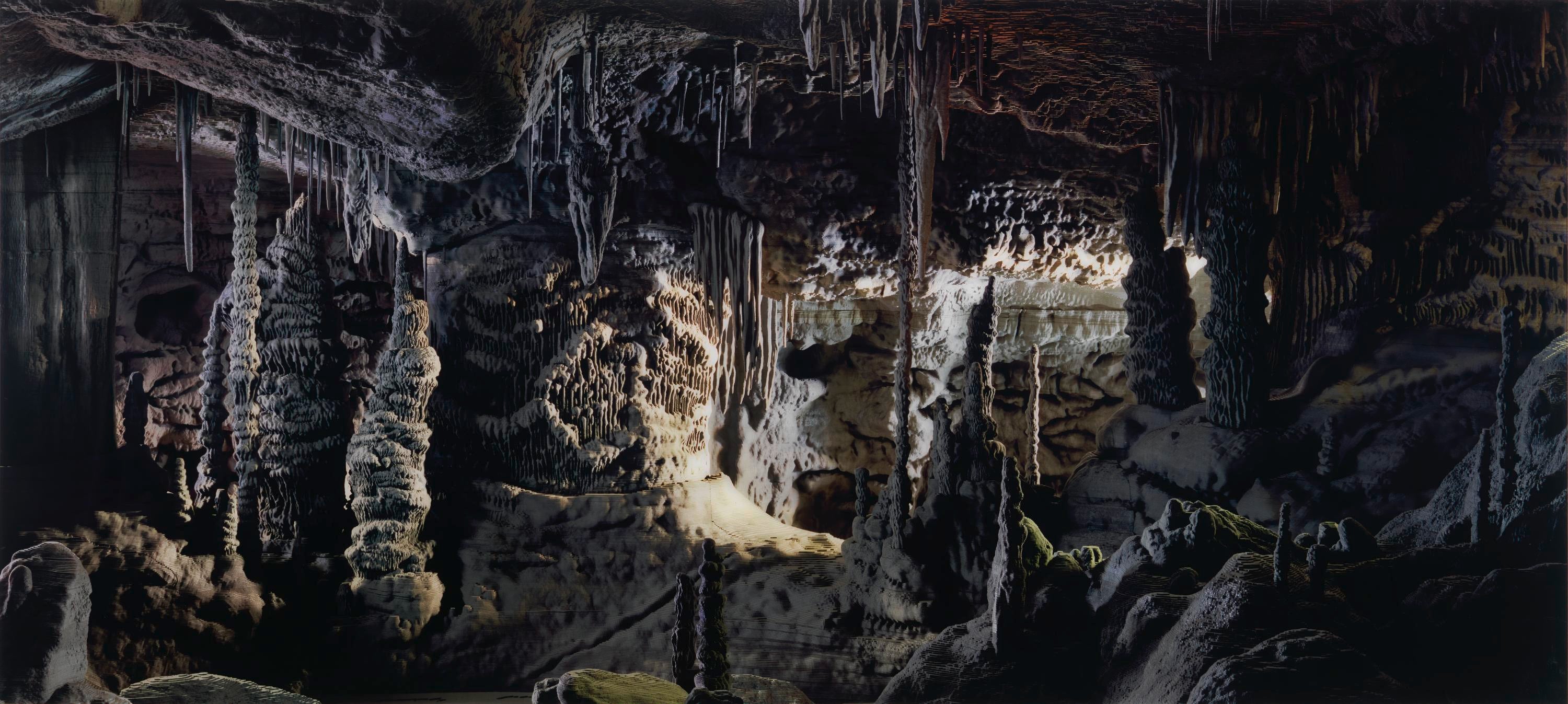 Cave interior in shades of green, grey, black and tan. Stalactites and stalagmites on view. Light entering from undefined space in background.