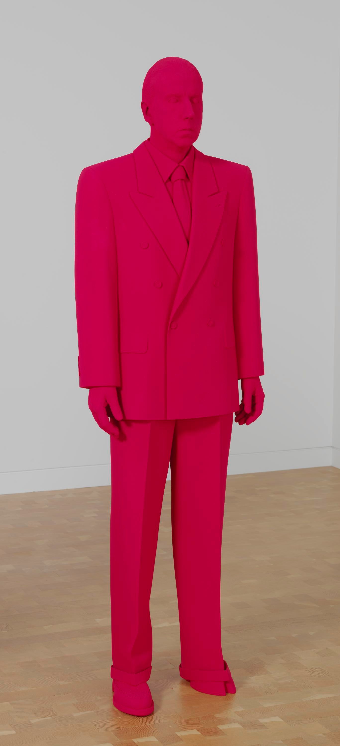 A monochrome red man stands in a room dressed in a red suit, red shirt, red tie and red shoes. One of his feet is a cloven hoof.