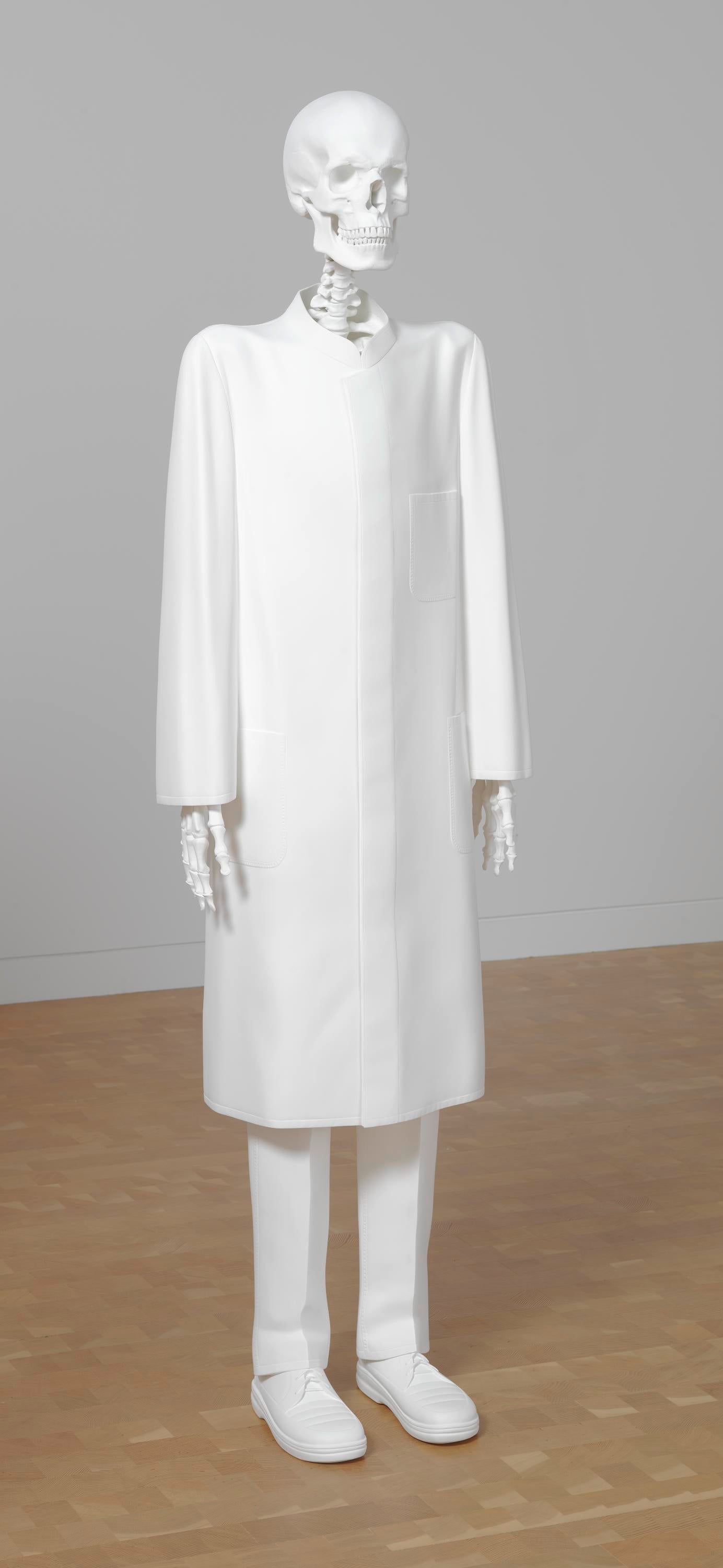 A white skeleton stands in a gallery space. It is wearing a lab coat, slacks, and shoes.