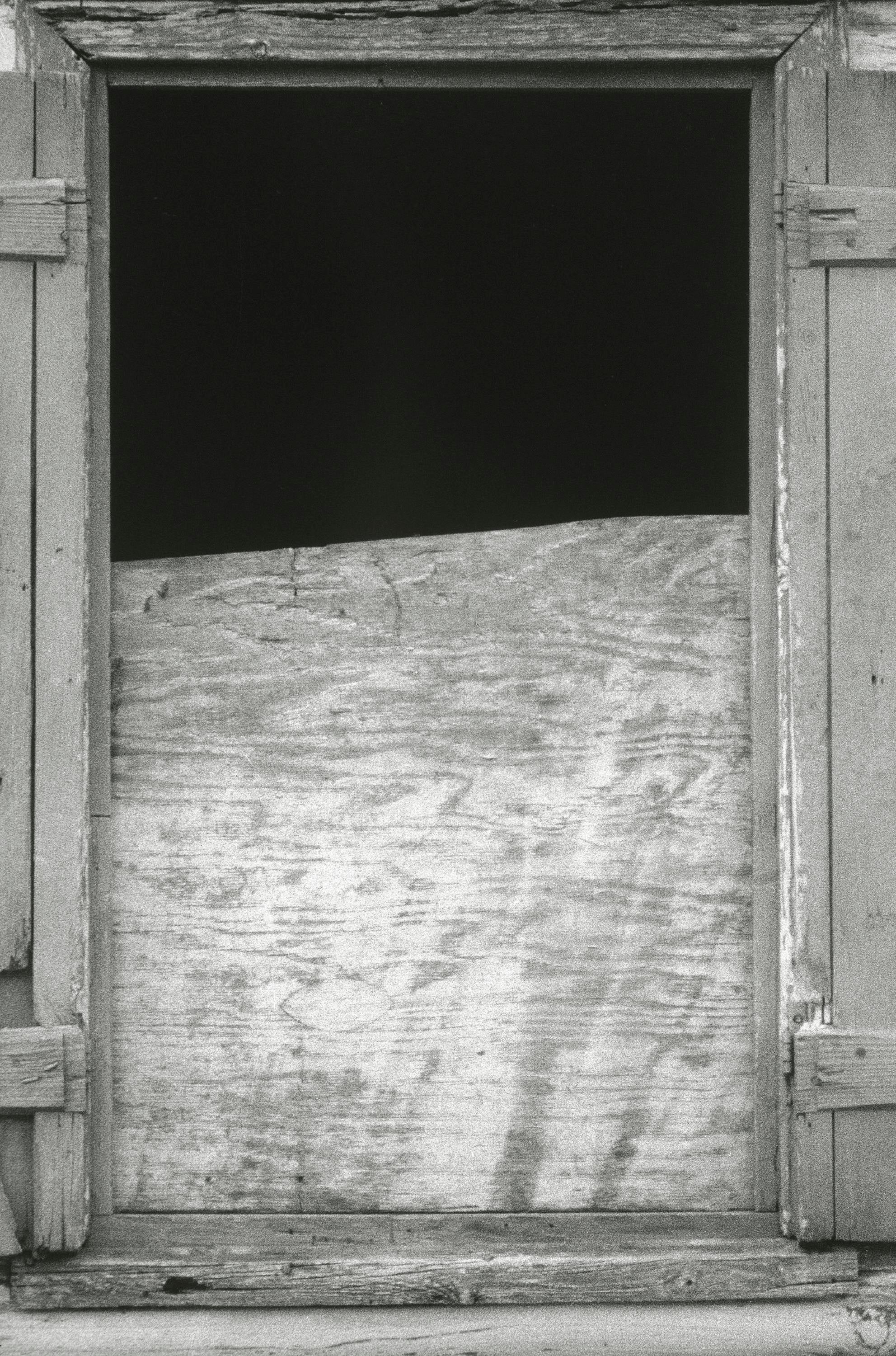 Una ventana parcialmente tapiada tiene un hueco negro en la parte superior donde la tabla está rota.