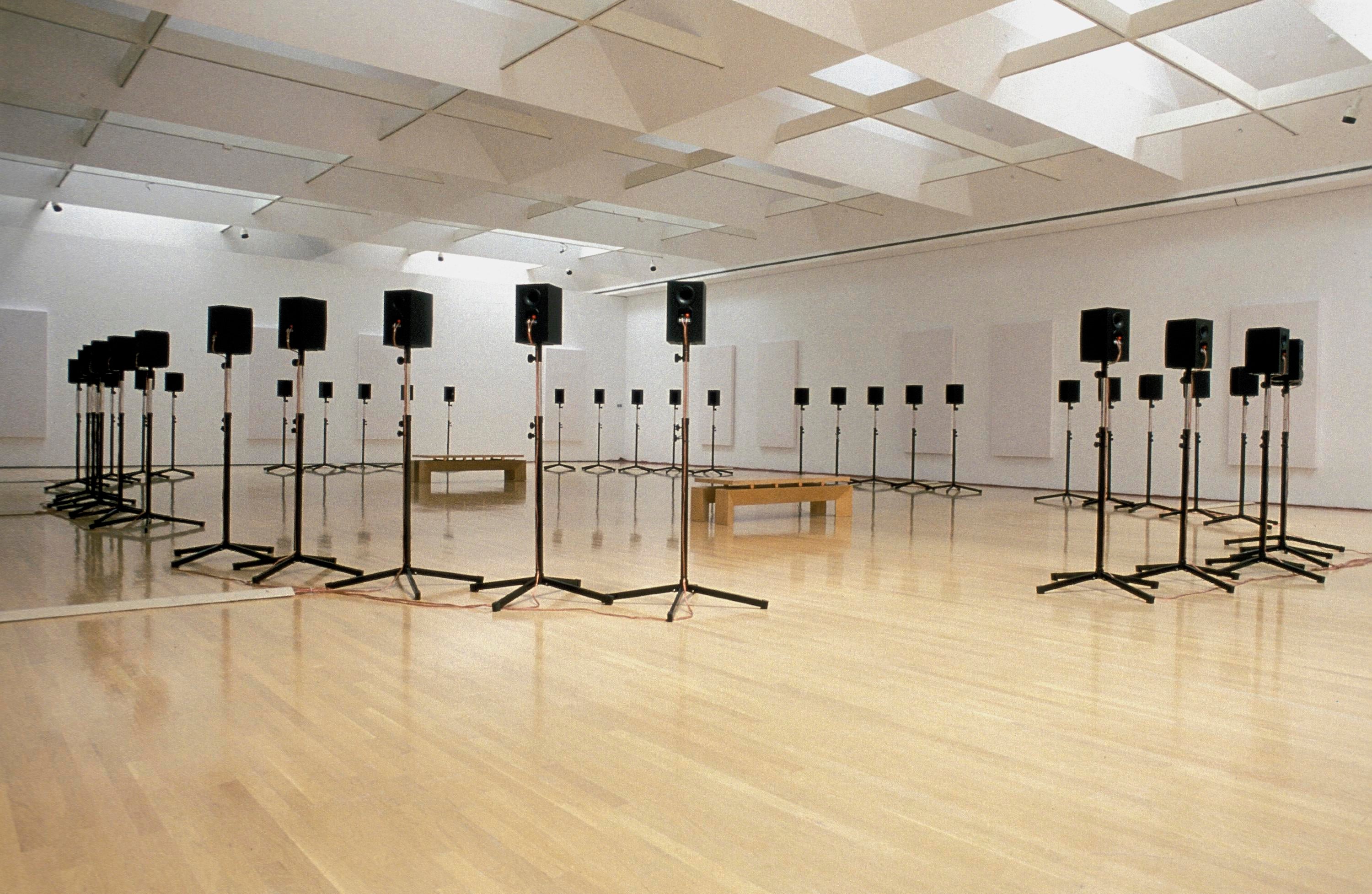 In a large white room with light wood flooring, a circle of black speakers on tripods surround two wooden benches.