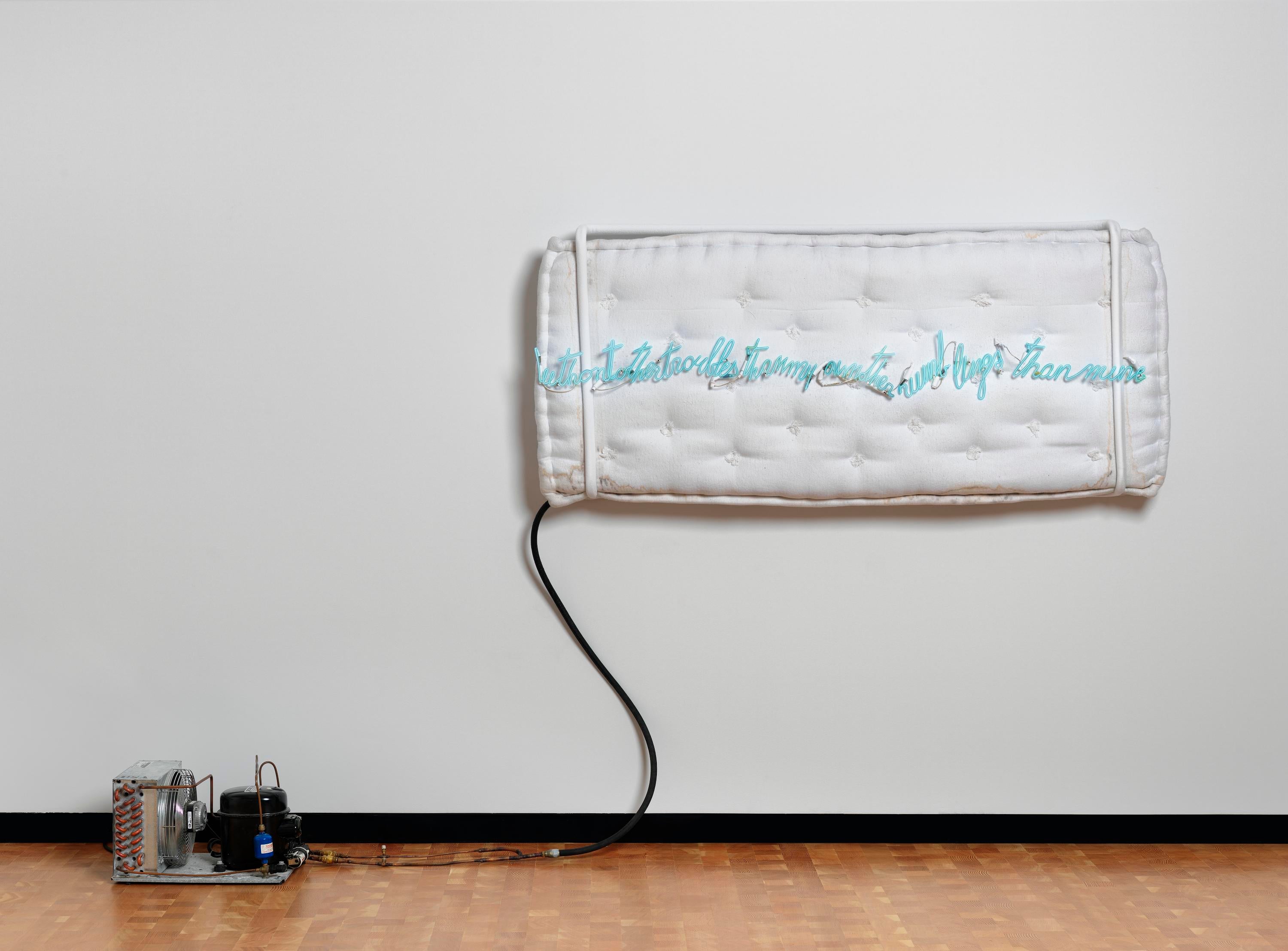 A mattress on a wall with blue neon tubing plugged into a transformer. The cursive neon says "without other troubles than my own other rumblings than mine."