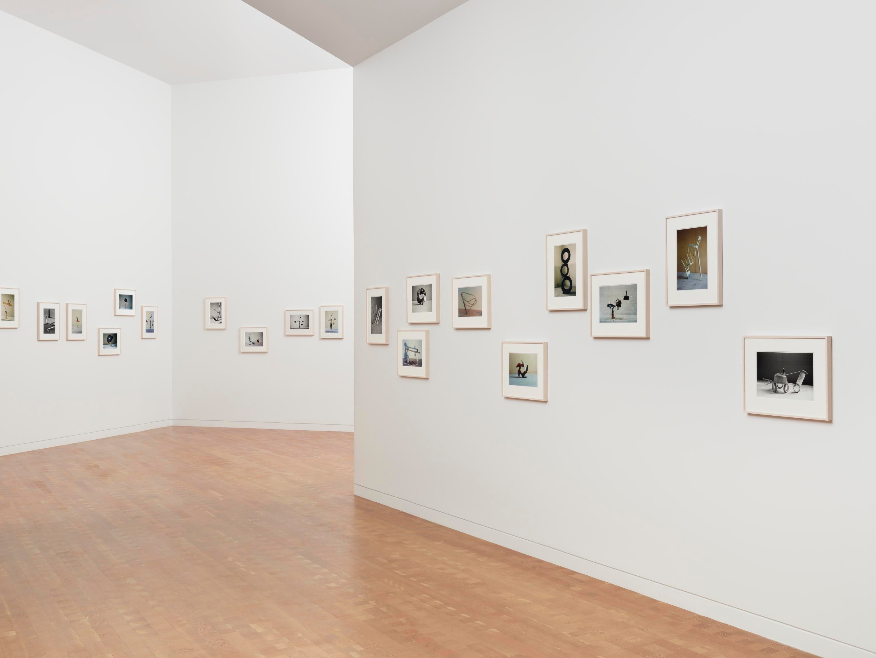 Nineteen framed photos hang on three different white walls, with a white ceiling and wooden floor.