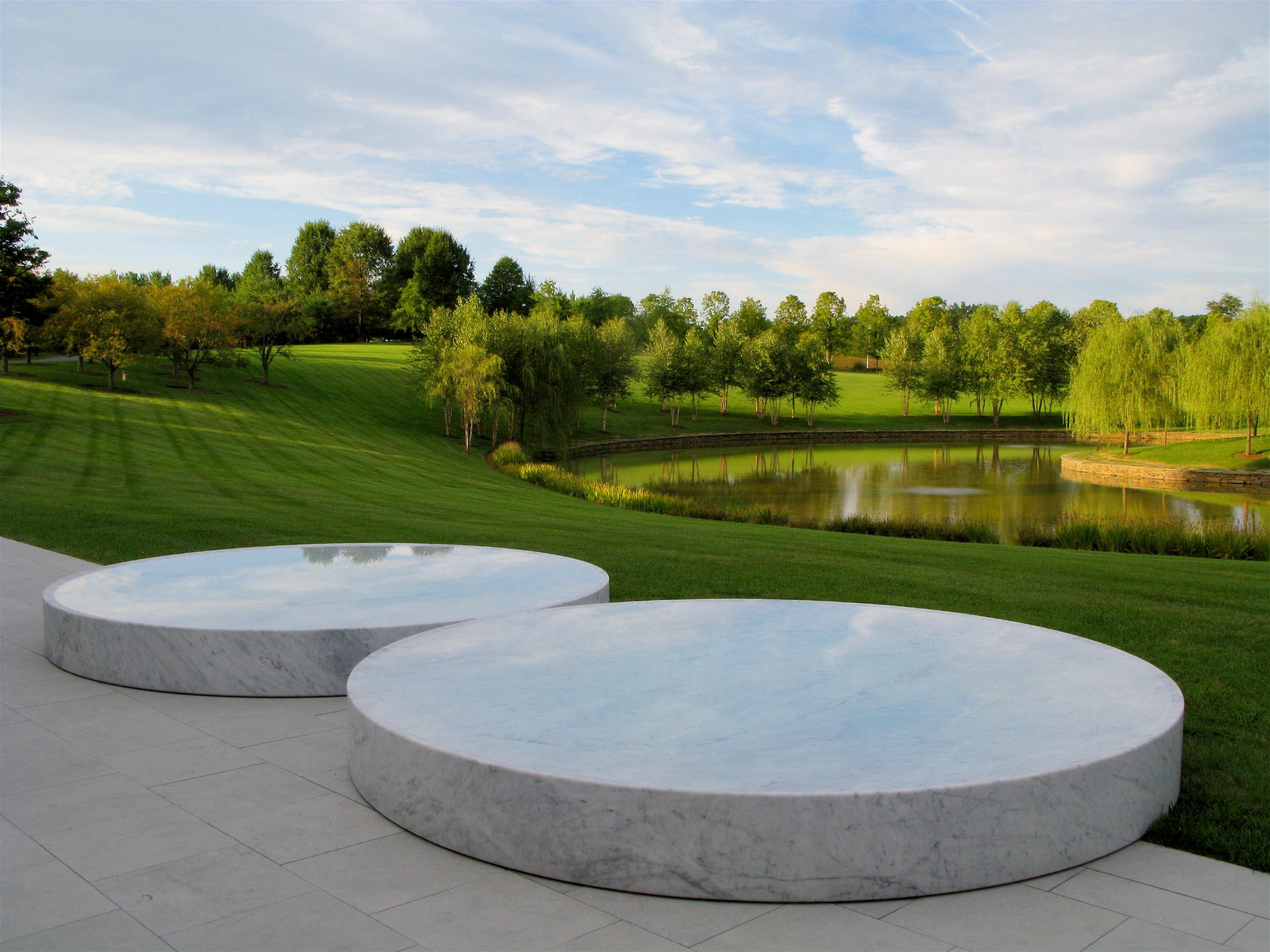 Dos piscinas circulares de mármol con agua se encuentran frente a un campo verde inclinado y un estanque, y árboles frondosos en el fondo.
