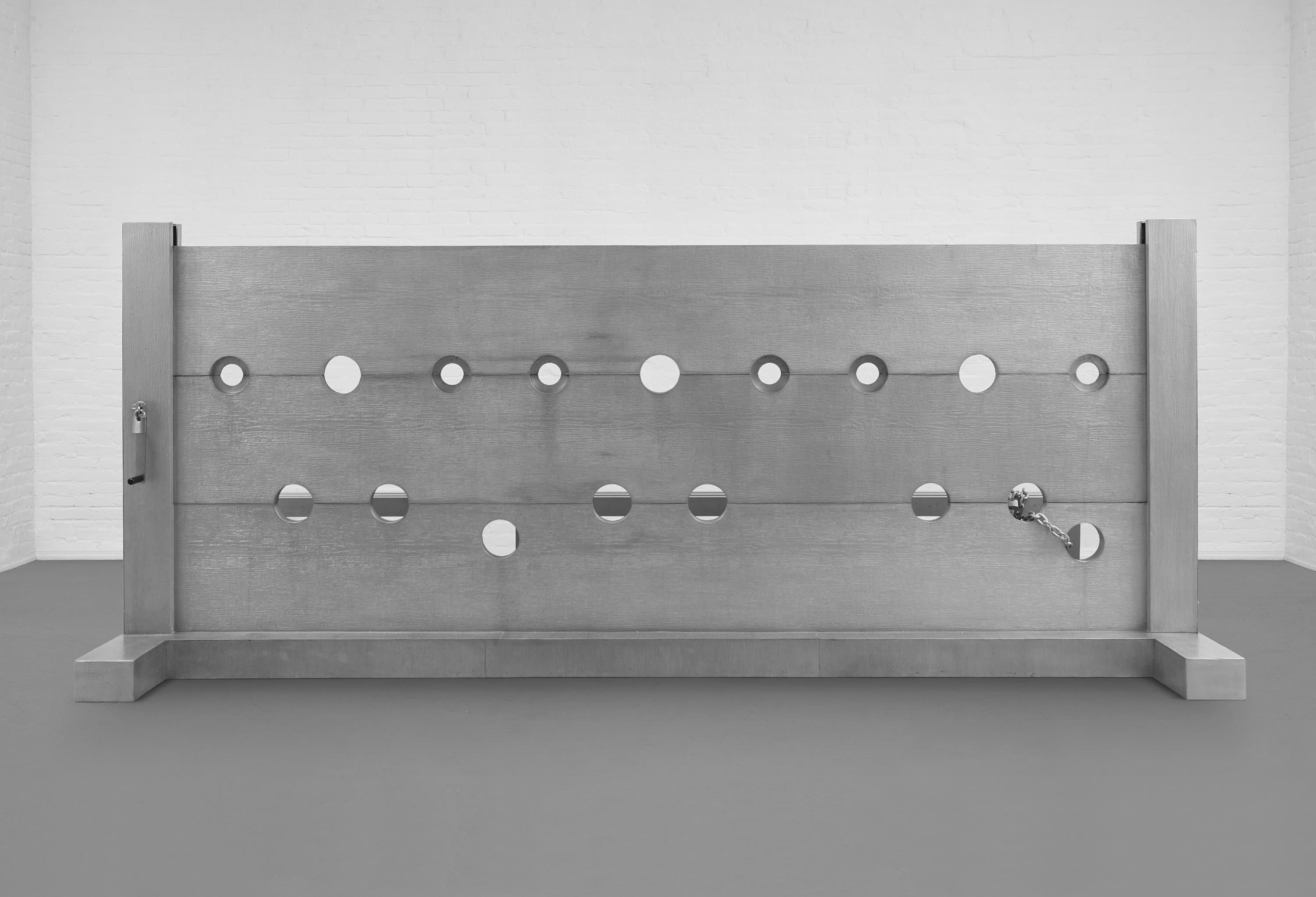 A large silver stockade sits on a grey floor in a white-walled room.