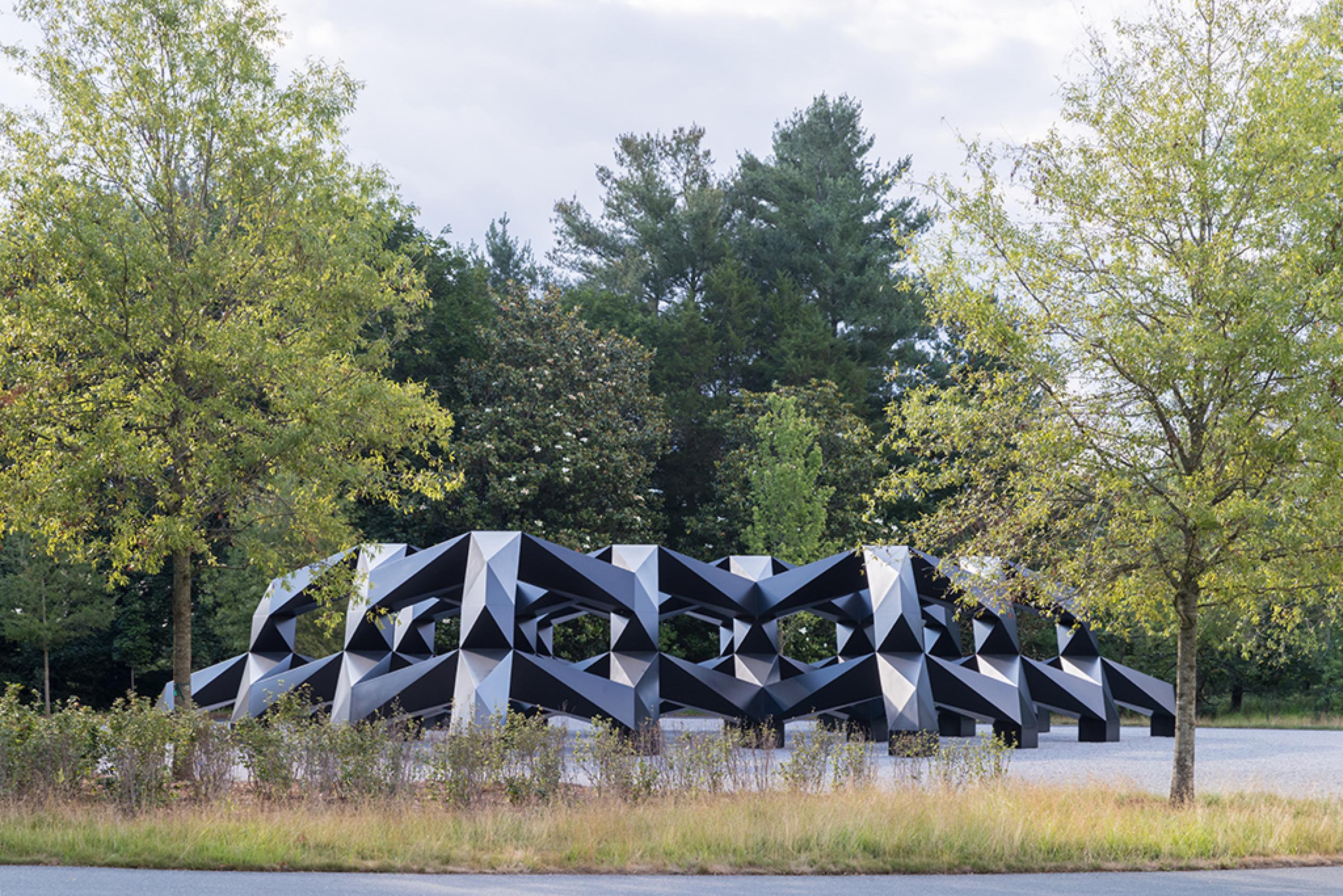 A large black outdoor sculpture comprised of geometric forms is surrounded by gravel and green bushes and trees.