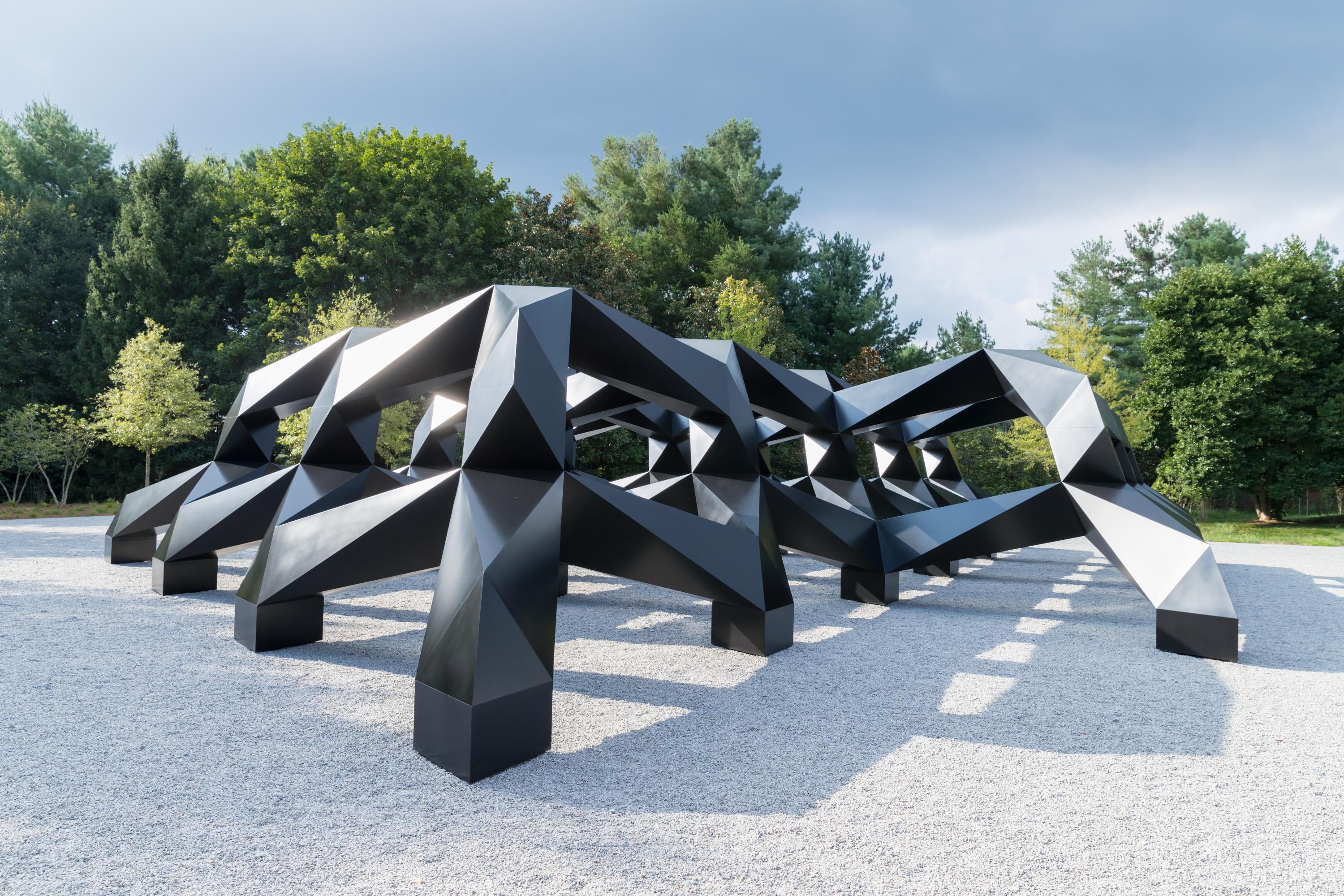 A large black outdoor sculpture comprised of geometric forms is surrounded by gravel and green bushes and trees.