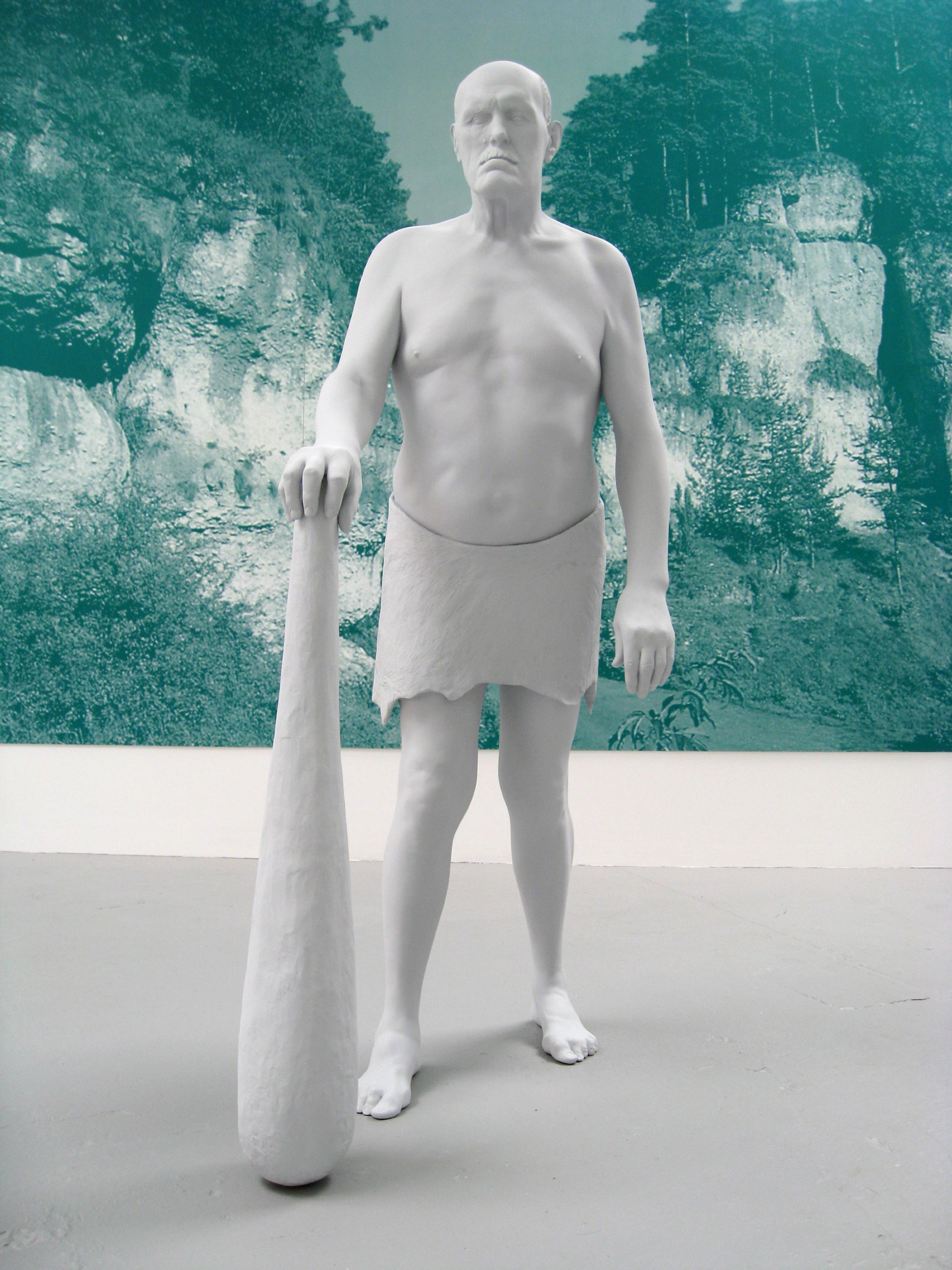 A giant old man naked from the waist up and dressed with a white rag from the waist down holds a white club. He is standing in front of a green and white silkscreen plastic panel with pictures of nature with rocks and trees