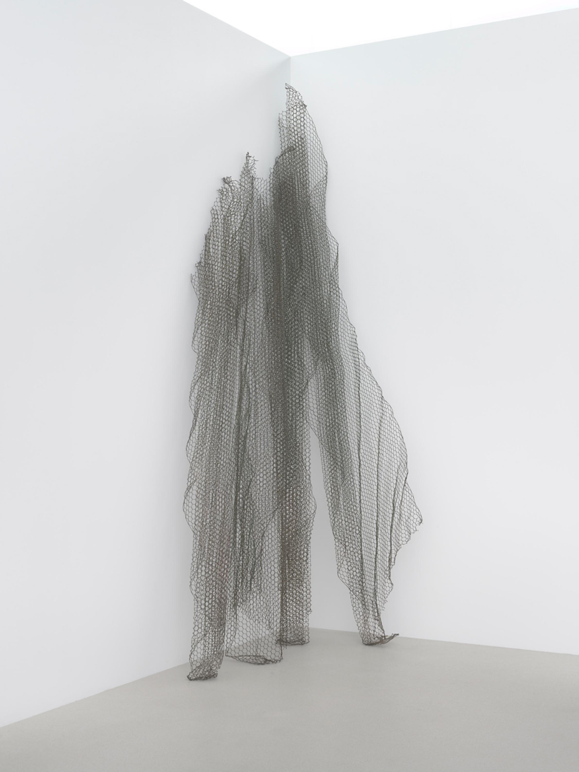 Four pieces of folded chickenwire lean against the corner of a white wall. They reach from the floor to the top of  the wall.