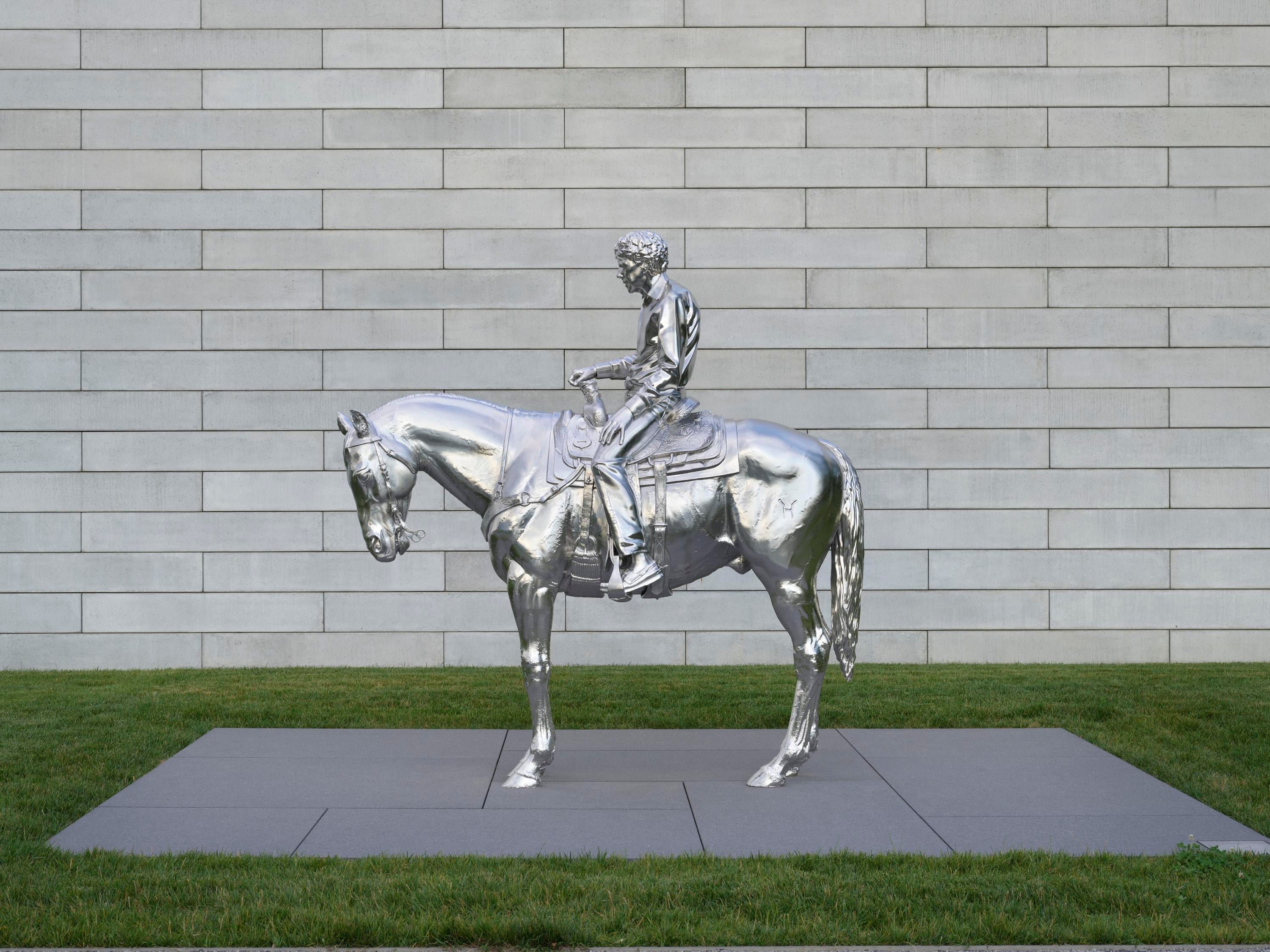 Una escultura de acero plateado representa a un hombre sentado sobre un caballo con una silla de montar. Detrás de la escultura, un muro de hormigón sirve de fondo, mientras que a su alrededor se extiende un campo de hierba verde.