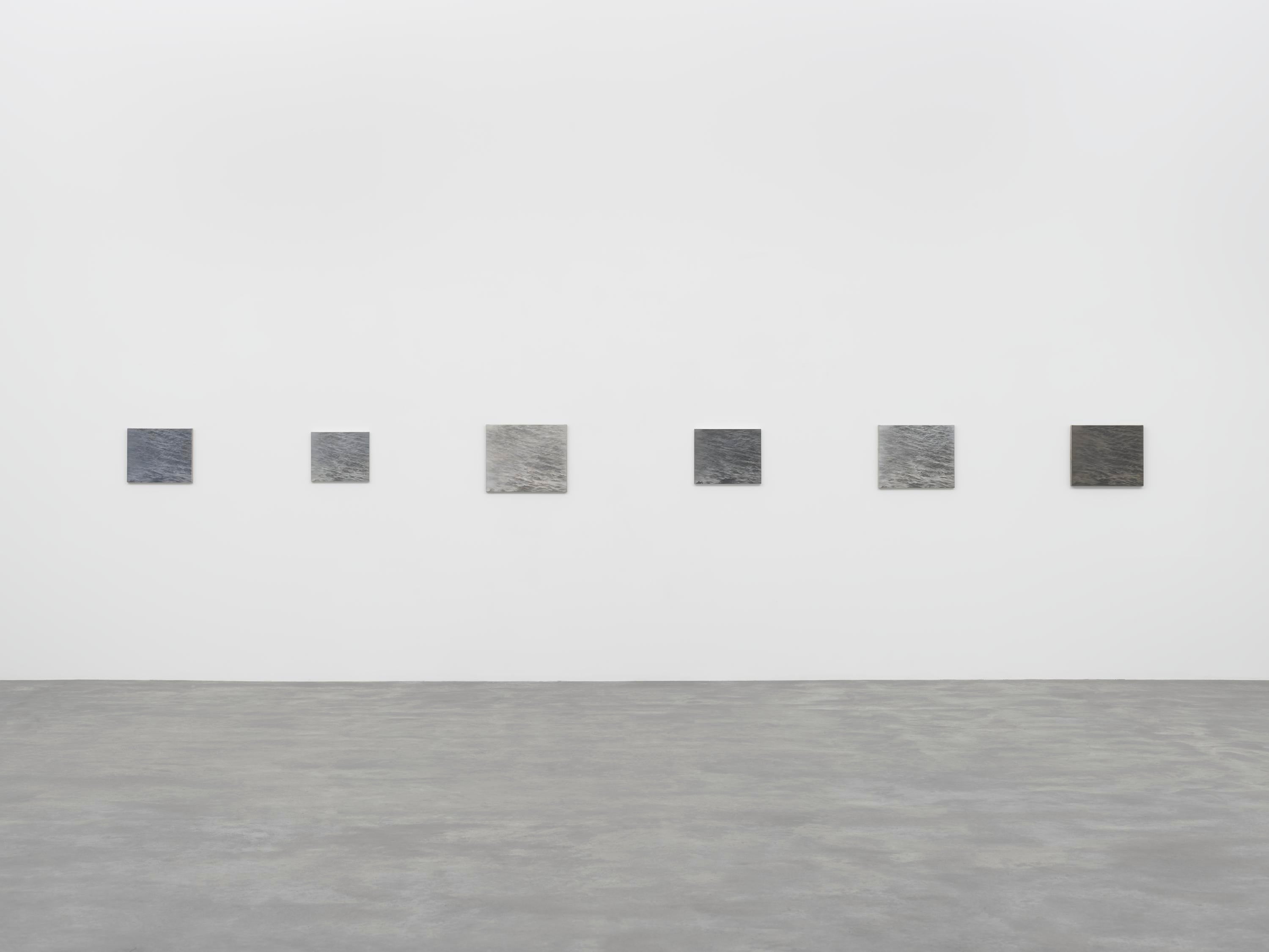 Una serie de seis lienzos grises cuelgan de una pared en una galería con suelo de piedra.