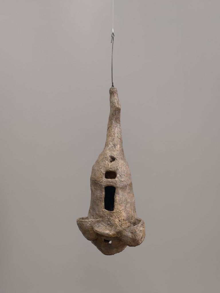 Una escultura de yeso (recubierta de goma laca) cuelga del techo a la altura de los ojos por medio de un gancho y un cable La escultura, que tiene una superficie irregular, presenta aberturas de varios tamaños en la superficie y proyecciones de elementos más allá de la superficie.