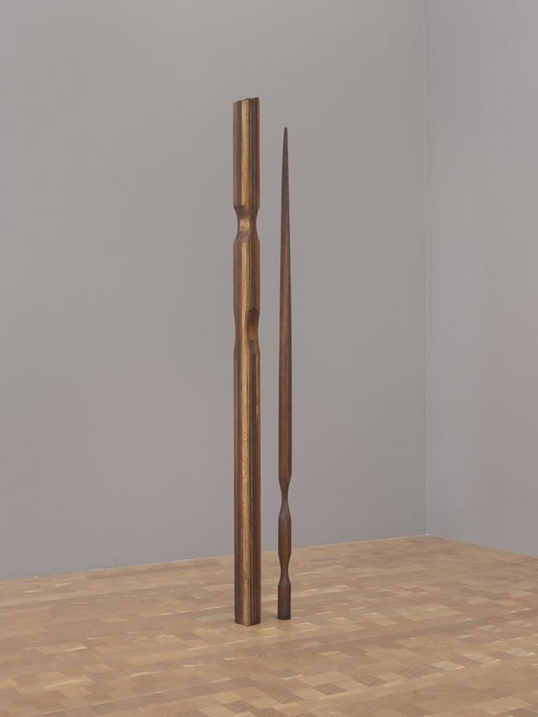 Dos formas altas y delgadas de madera torneada y tallada, una que se asemeja a un poste de cama y otra que se asemeja a una lanza, se ubican en el suelo de la galería.