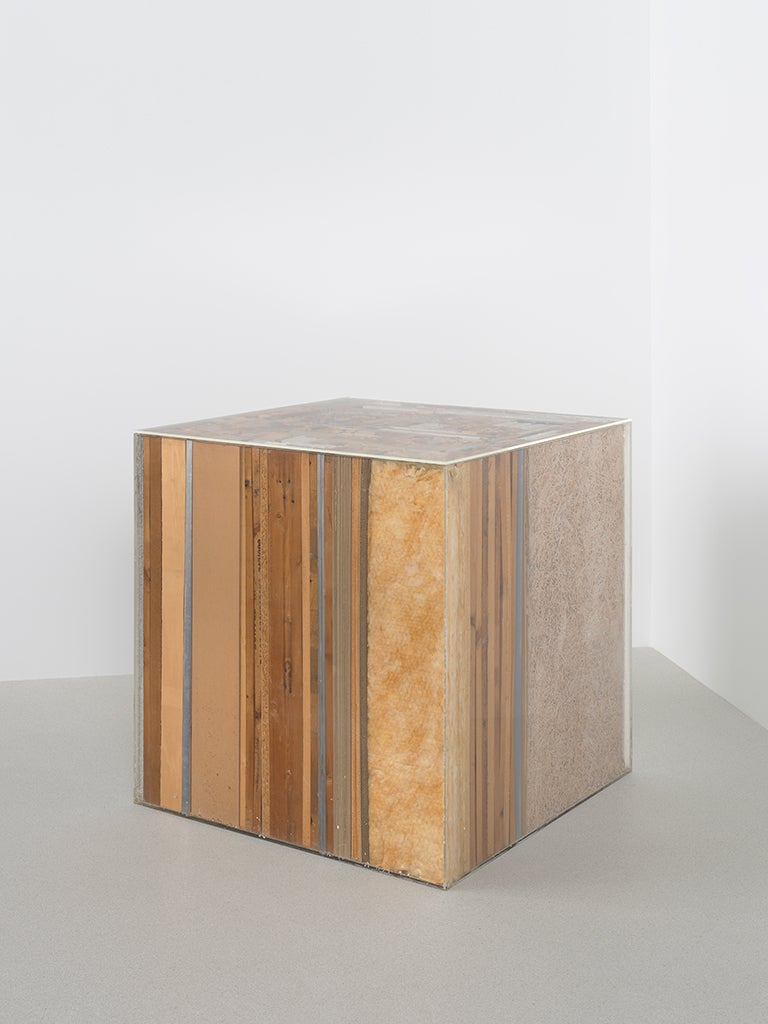 A plexiglass cube sits on a gallery floor, and it is full of wood, insulation, and various other materials.