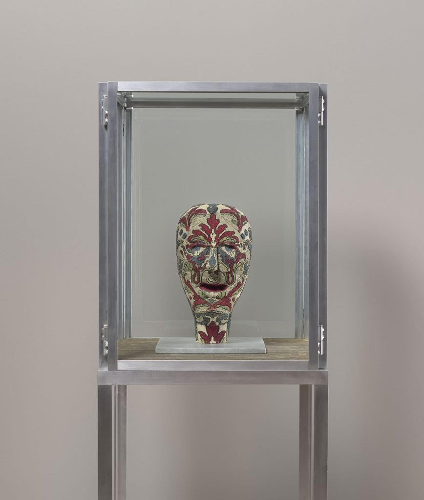 En el interior de un armario de aluminio metálico montado en el suelo descansa una cabeza figurativa esculpida hecha de tela puntiaguda multicolor que se apoya sobre una plataforma.