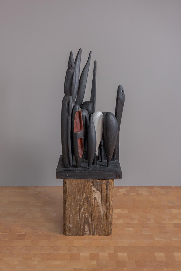 Sobre un suelo de madera, frente a una pared pintada de gris, se ubica un pedestal de madera toscamente tallado que sostiene una escultura abstracta de madera pintada de color negro, con toques de rojo y blanco.