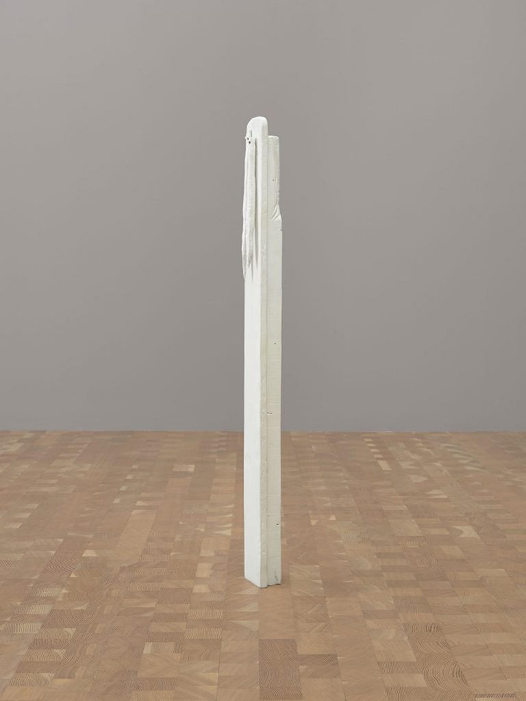 Frente a una pared gris, una escultura de madera alta y delgada con figuras blancas se encuentra directamente sujeta al piso de madera.