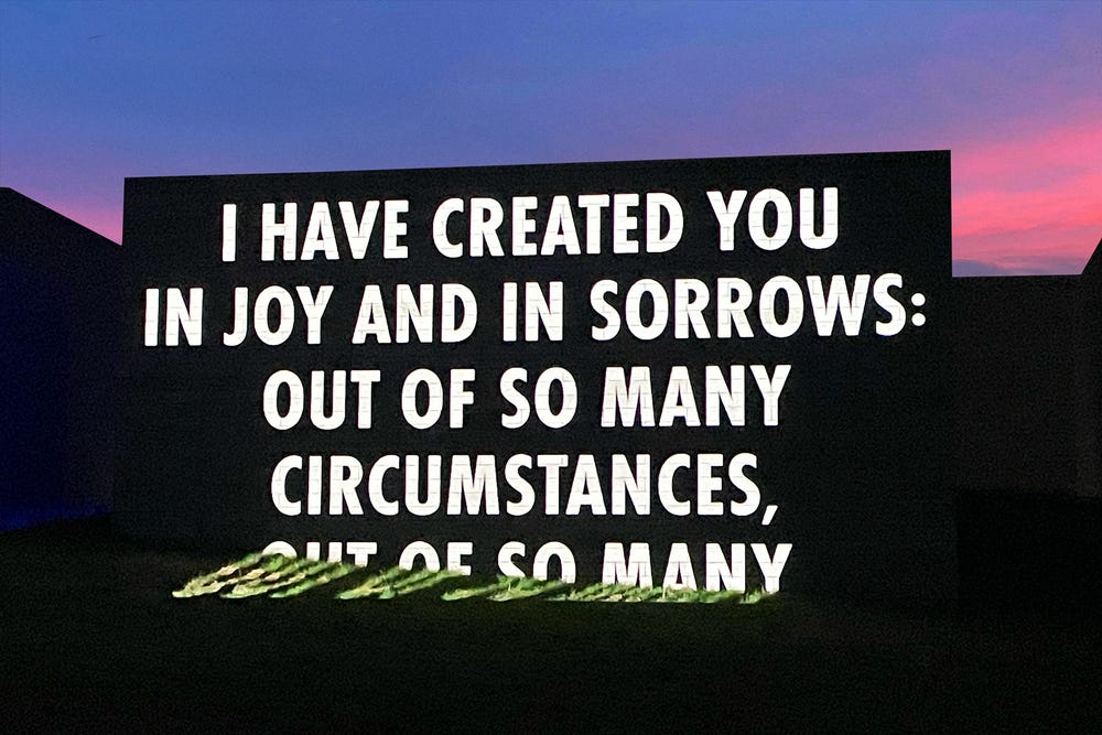 Text projected on a building at sunset that says I HAVE CREATED YOU IN JOY AND IN SORROWS: OUT OF SO MANY CIRCUMSTANCES,