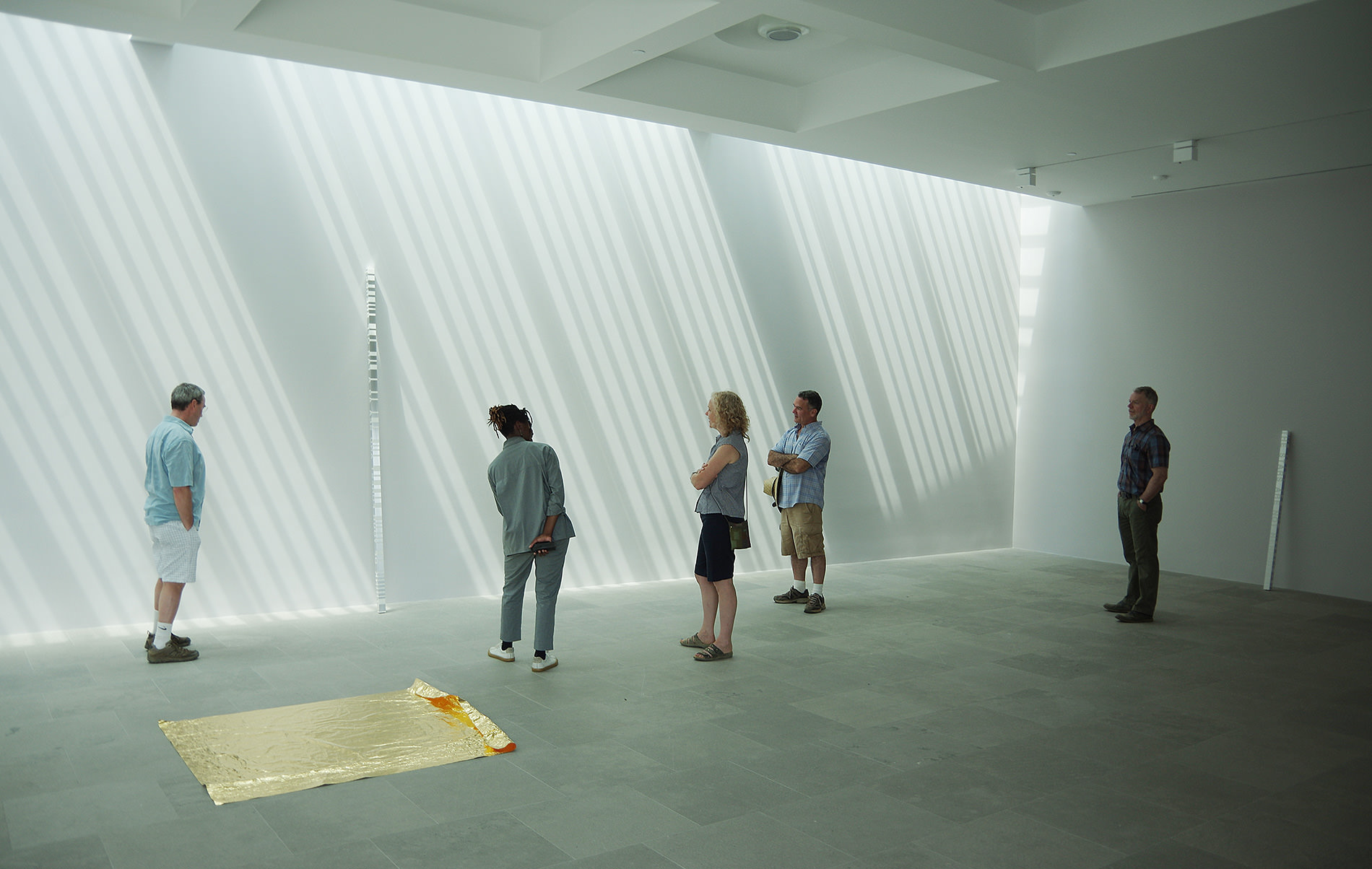 Five people, one dessed all in grey, converse in a room with white walls and gray stone floor, near gold and translucent sculptures.