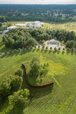 An outdoor aerial view of a metal sculpture winding through a field in front of a large geometric sculptures and a tree-lined path. Beyond the path is a cluster of minimal concrete buildings surrounded by trees.