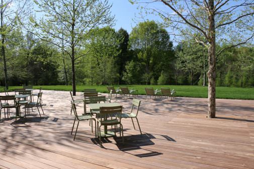 Chairs and tables sit on a wooden deckat the edge of a lawn with a forest in the background.