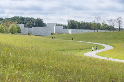 A gravel path cuts through an expanse of meadow. A number of visitors are moving toward the grey concrete buildings in the distance. Trees amass further in the landscape.