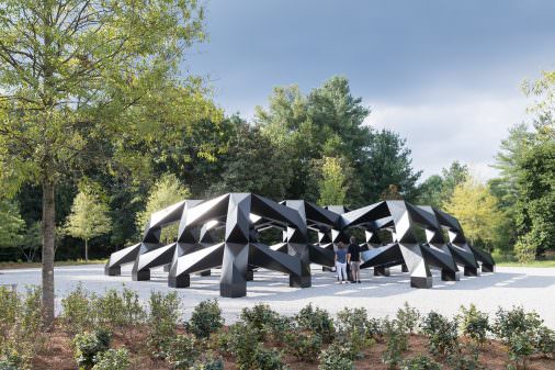 Two people look at a large black geometric sculpture comprised of triangular forms, surrounded by gravel and green bushes and trees.