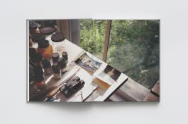 A book sits on a white surface, and it is open to a full-page spread photograph of Brice Marden's Studio. Pigments and paints and photographs sit on the table.